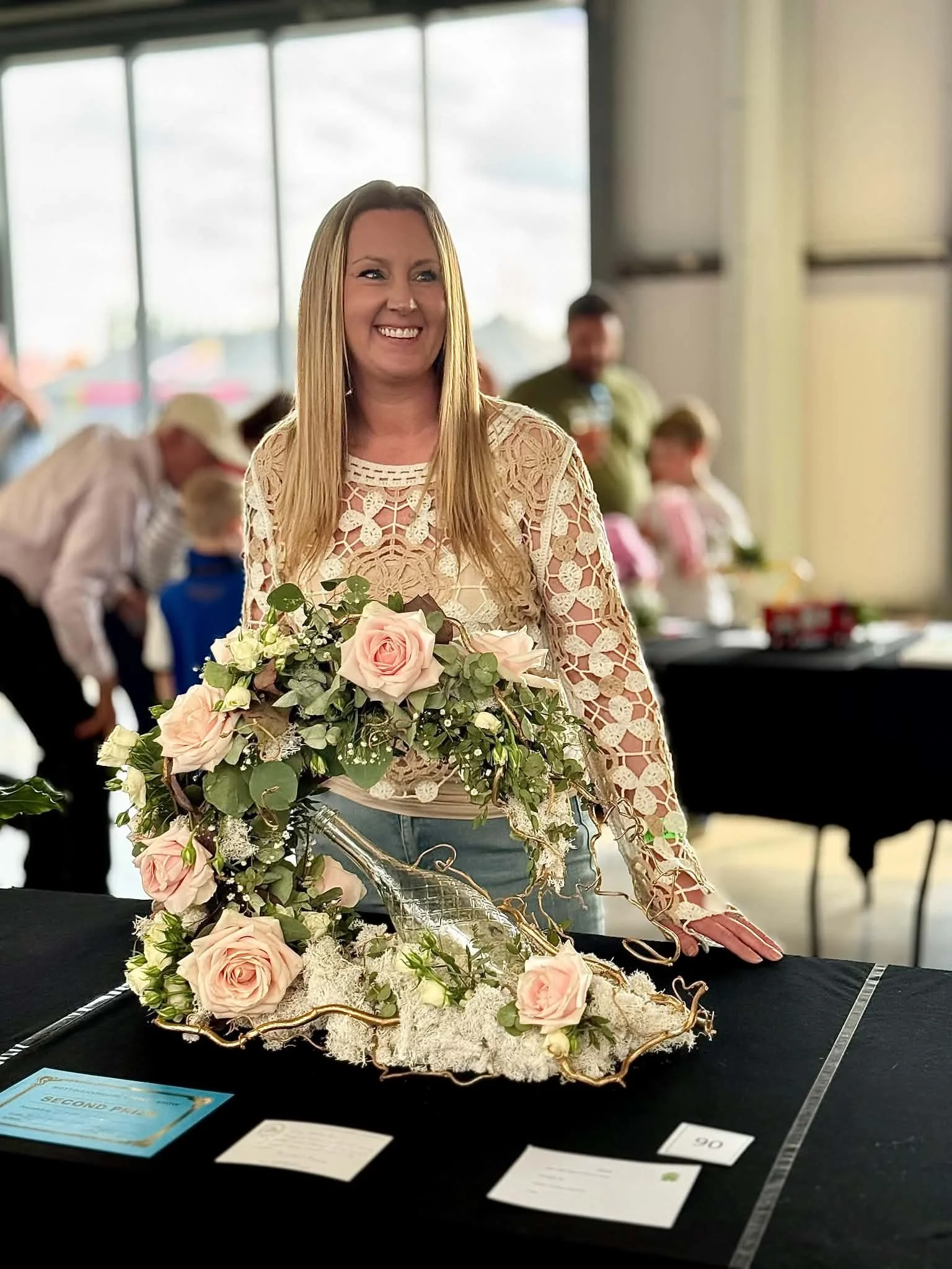 A woman with long blonde hair, wearing a lace top, holding a bouquet of pink and white roses and greenery, smiling at a flower arrangement displayed on a black table in a bright indoor space with large windows, with people in the background.