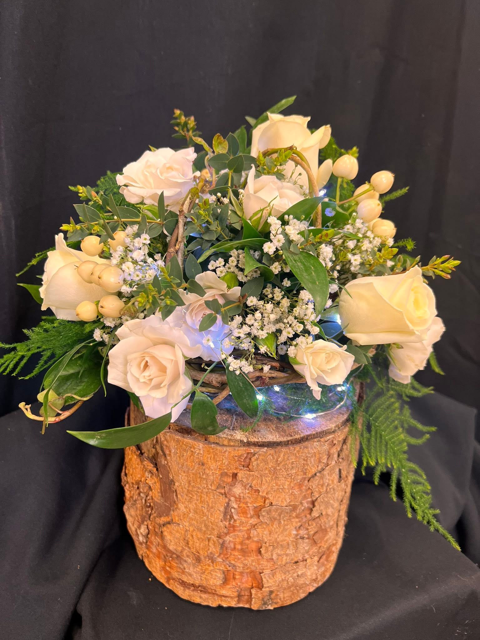 A floral arrangement with white roses, small white flowers, green leaves, and berries in a glass vase placed on a tree trunk against a black background.