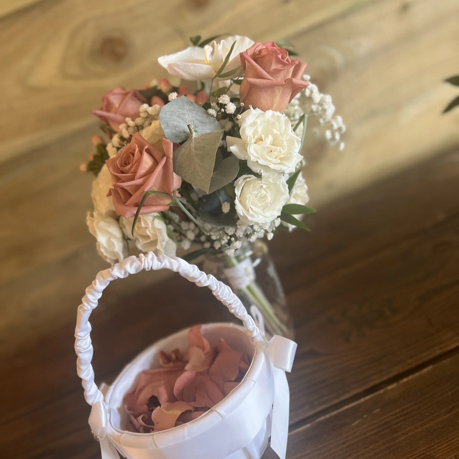 Close-up view of a flower arrangement with pink roses, white flowers, and greenery in a white basket with a ribbon, placed on a wooden surface.