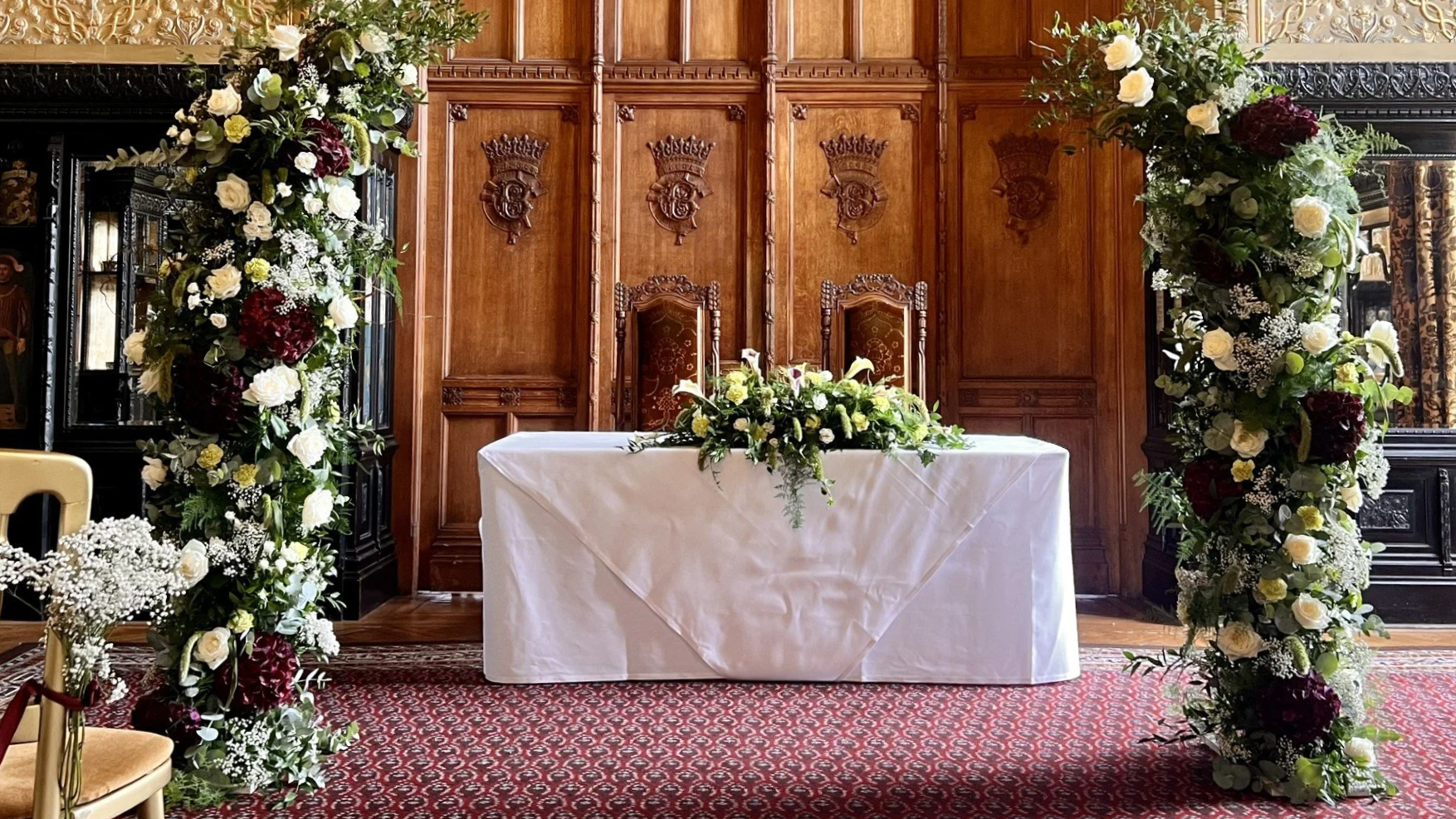 A wedding ceremony setup with a table covered in a white cloth, adorned with a floral centerpiece, framed by two tall floral arrangements of white, purple, and green flowers, against a wooden paneled wall with carved crest decorations.
