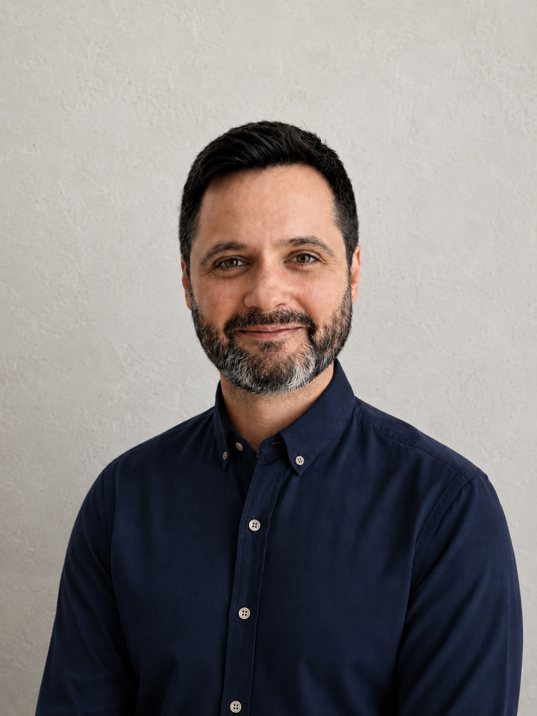 A man with dark hair and a beard, wearing a navy blue button-up shirt, standing against a beige wall.