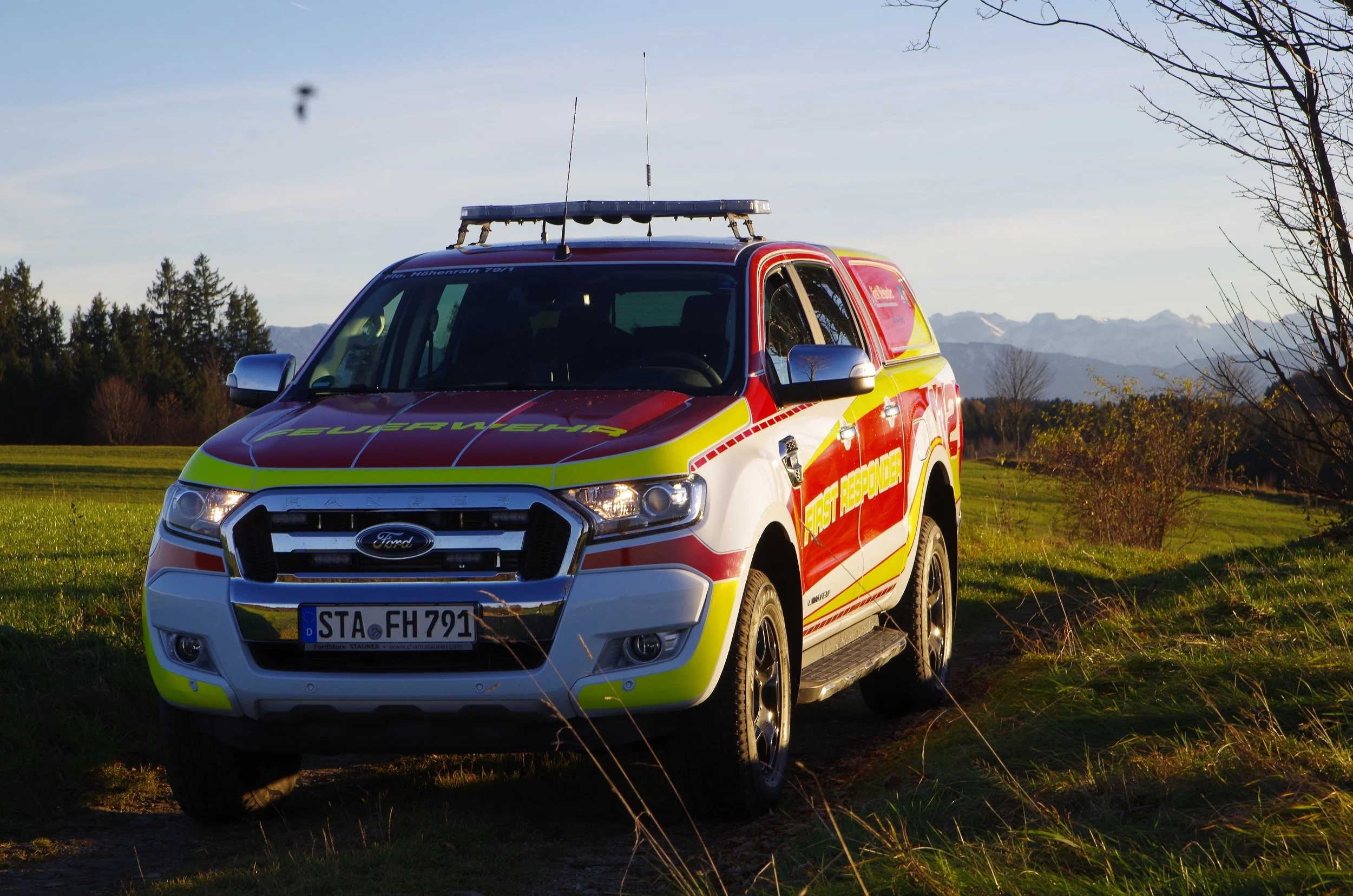 Das Einsatzfahrzeug vor einer ländlichen Kulisse mit Bäumen, Wiesen und Bergen im Hintergrund, Sonne scheint, Himmel ist blau.