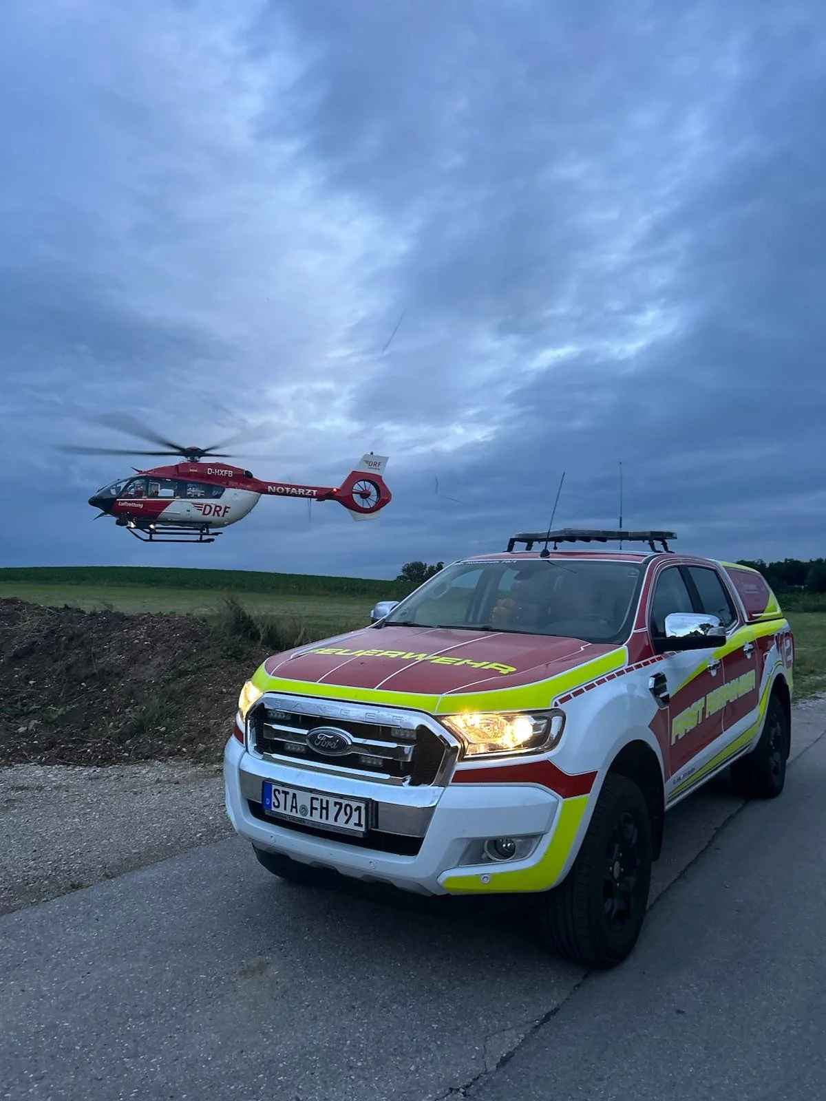Das Einsatzfahrzeug und ein Hubschrauber in einer ländlichen Umgebung bei Dämmerung, mit einem bewölkten Himmel im Hintergrund.
