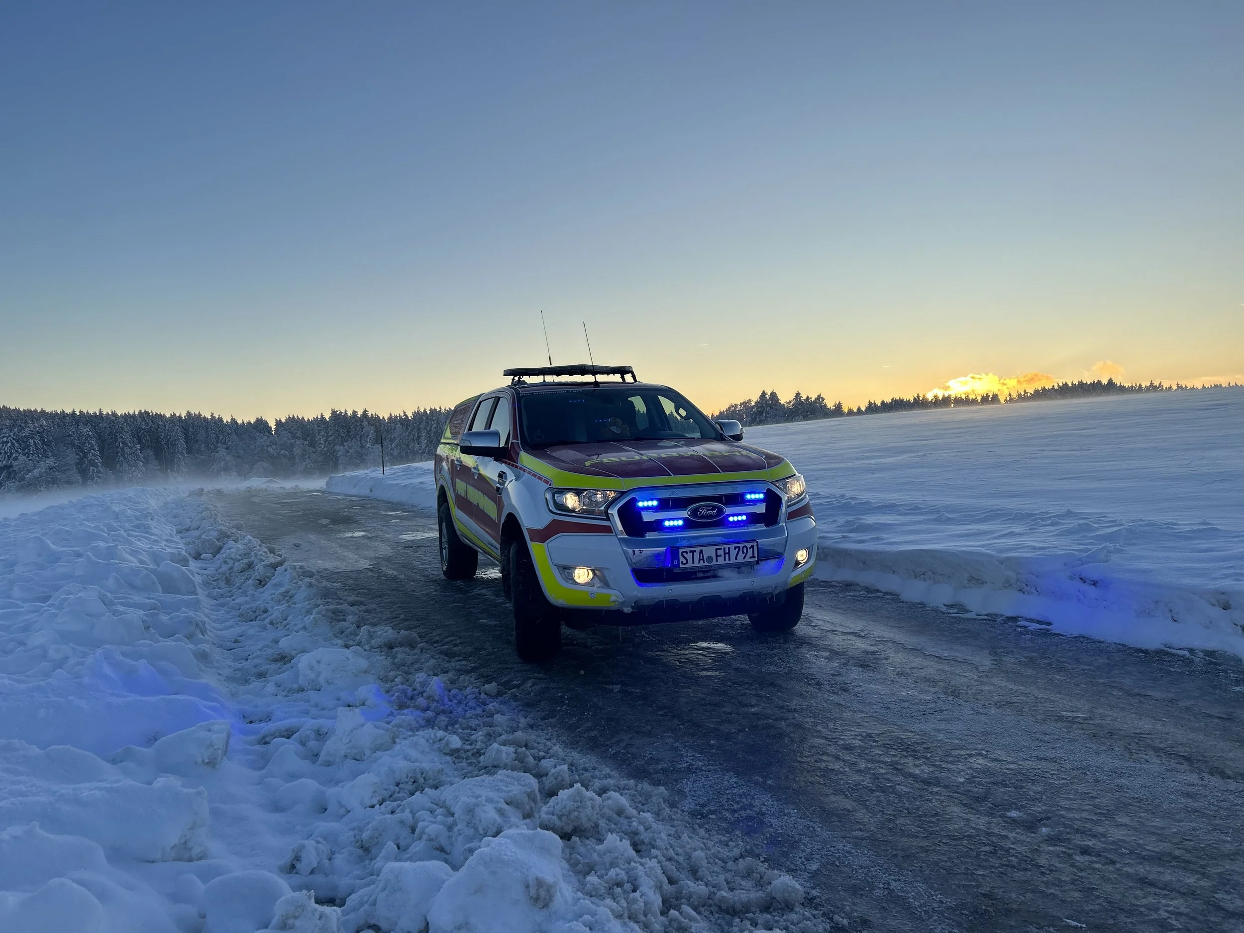 Das Einsatzfahrzeug auf schneebecktem Feld bei Sonnenuntergang, mit Blinklichtern eingeschaltet.
