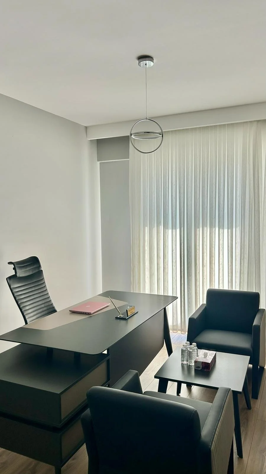 Office with modern furniture, black leather chairs, a desk with a pen holder, water bottles on a side table, and curtains covering a window.