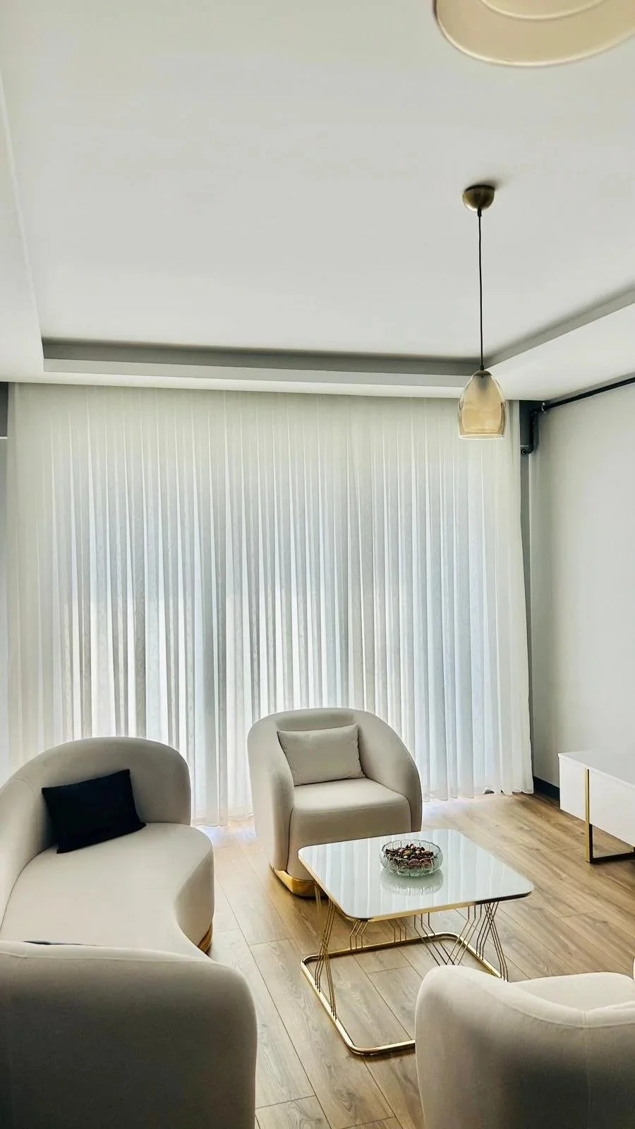 A modern living room with white curved sofas and a glass-top coffee table with gold legs. Light-colored curtains cover a large window, and there is a pendant light hanging from the ceiling.