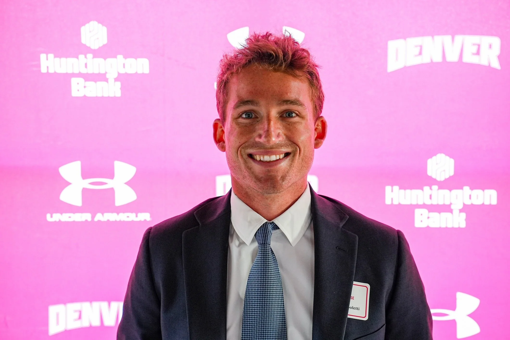 A smiling man in a suit and tie standing in front of a pink backdrop with logos and text that include Huntington Bank, Under Armour, and Denver.