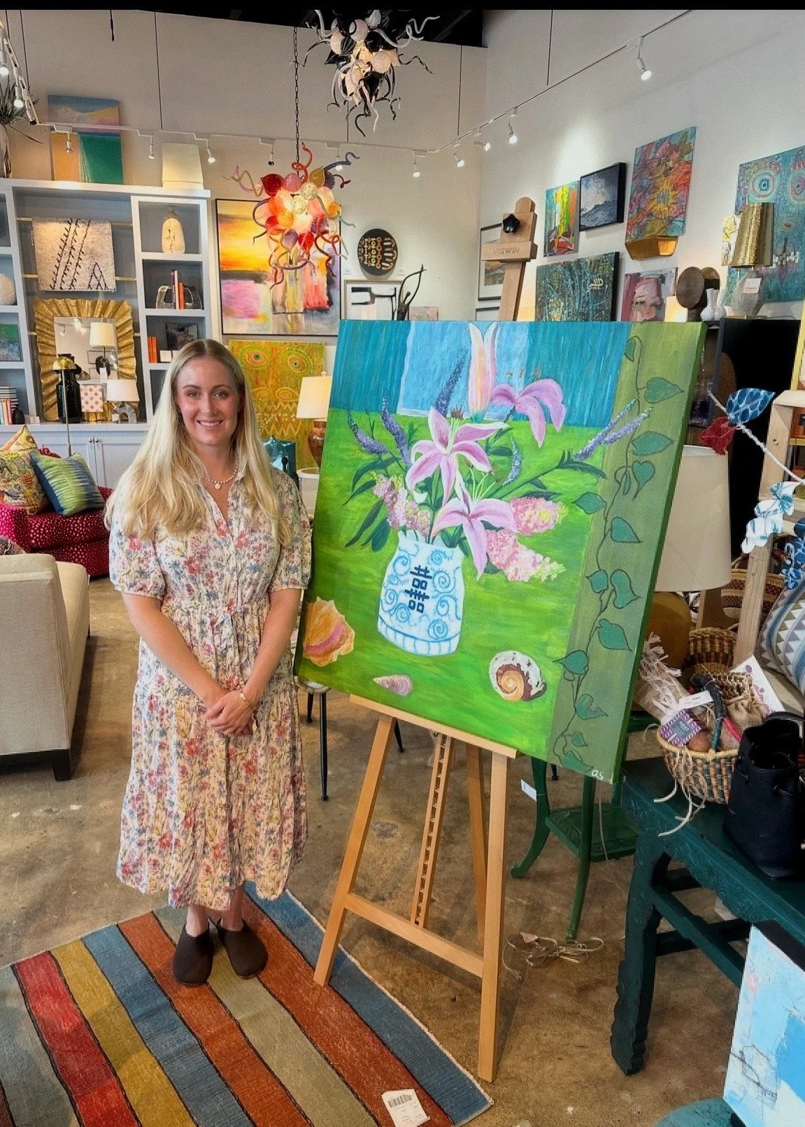 A woman with long blonde hair wearing a floral dress standing next to an easel with bright floral painting in an art gallery, surrounded by colorful art pieces and decor.