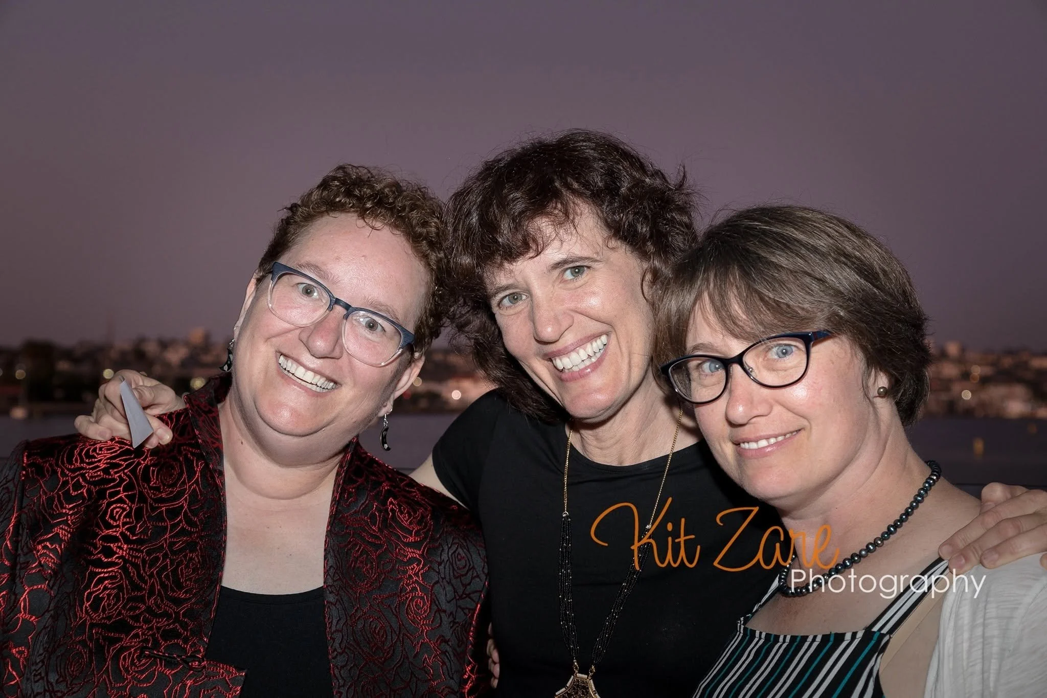 Group of three sisters smiling for a photo, with a cityscape in the background during evening.