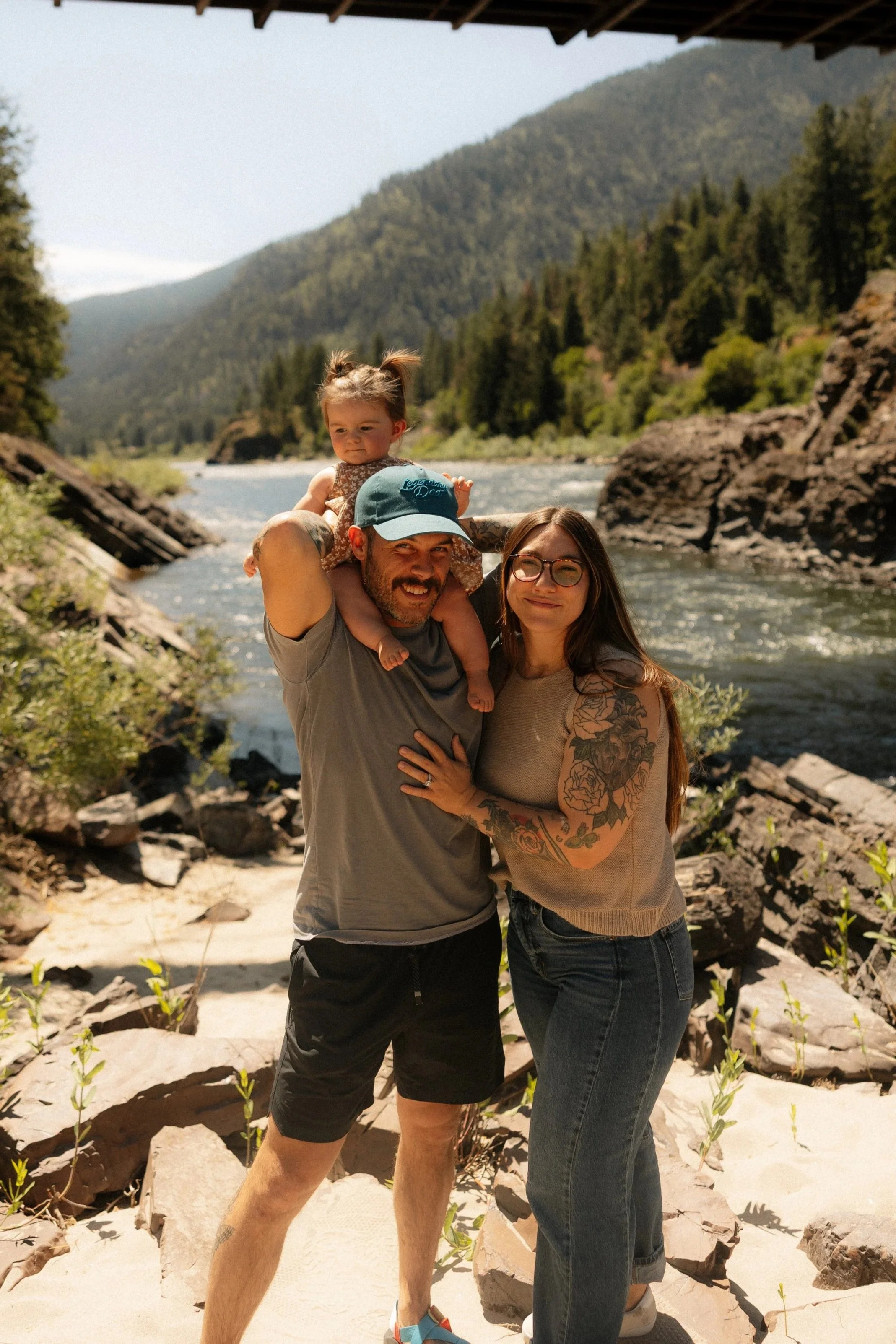 A family of three posing by a river in a mountainous area with trees in the background. The man is carrying a young girl on his shoulders, and a woman stands beside them.