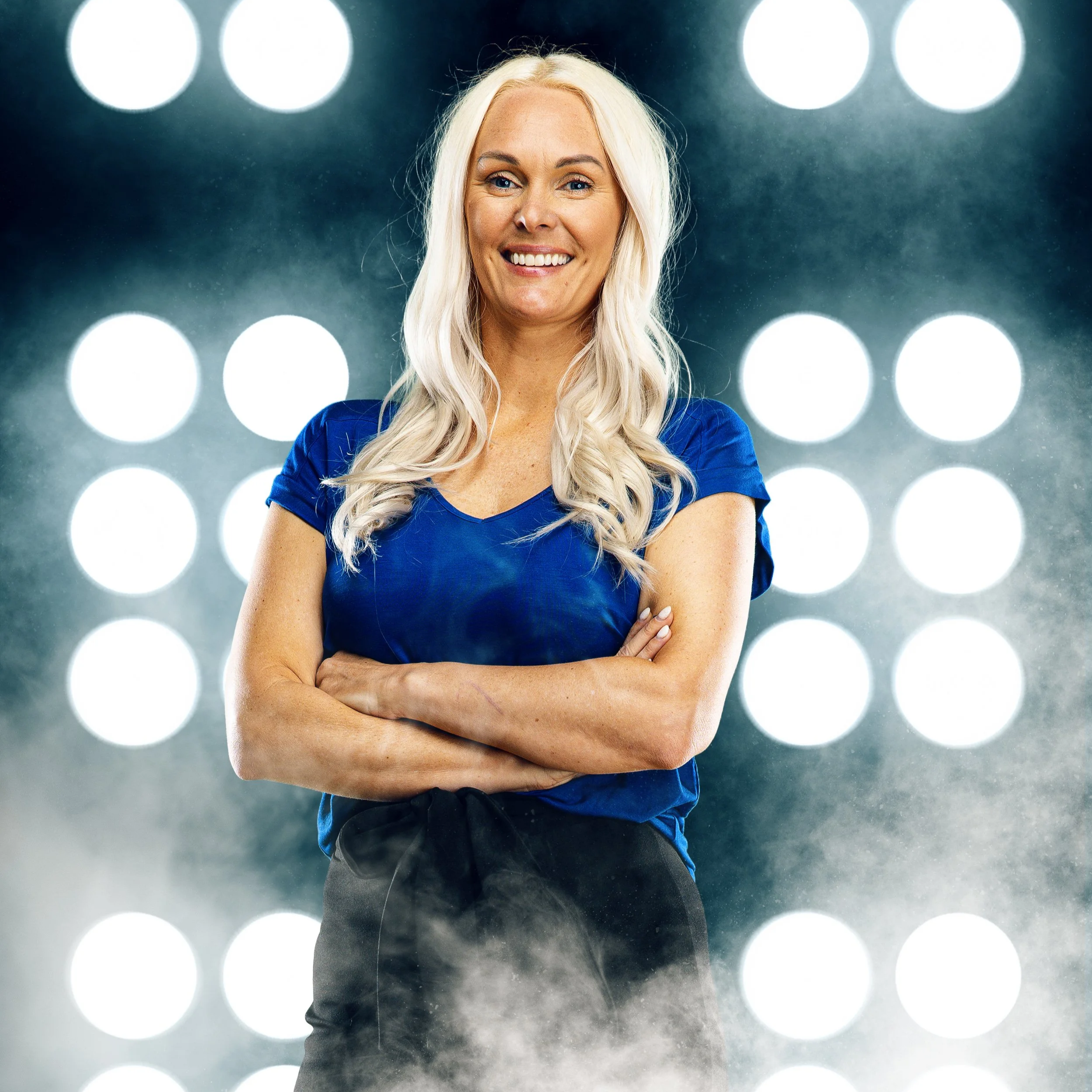 A woman with long blonde hair, smiling with her arms crossed, is standing in front of a background of large, illuminated white circles arranged in a grid pattern.