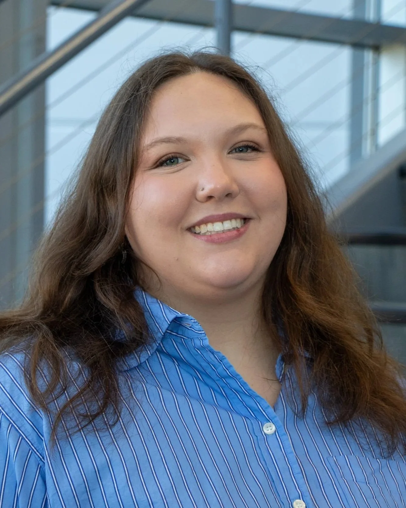 A woman with long brown hair, blue eyes, and a nose piercing smiling indoors near a staircase, wearing a blue striped button-up shirt.