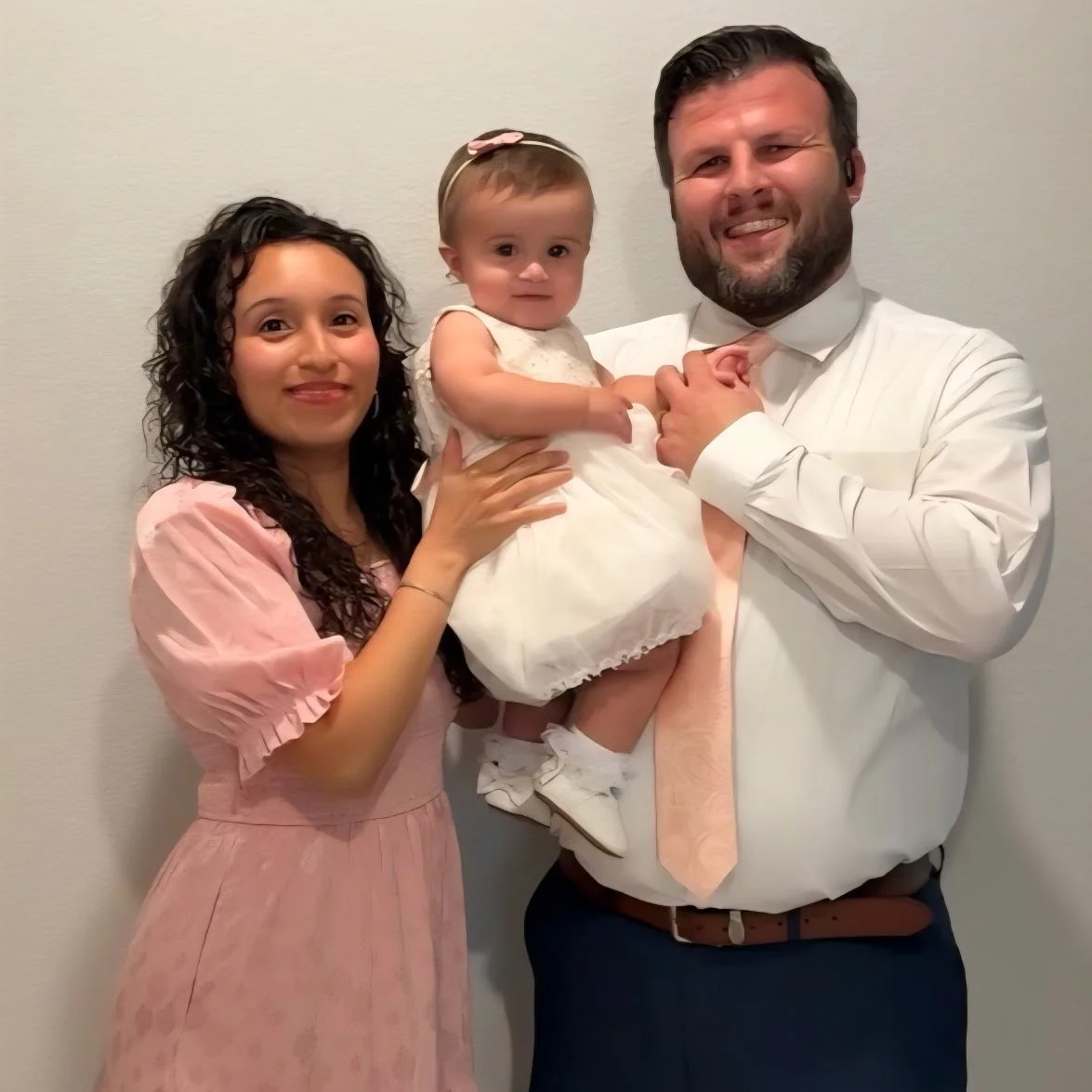 Family of three with a man, woman, and a baby girl dressed in formal attire, standing against a plain wall.