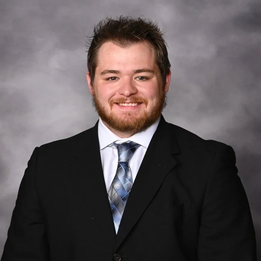 A man wearing a black suit, white shirt, and a plaid tie, smiling against a gray background.