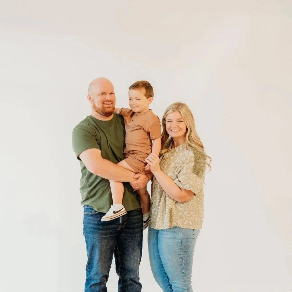 A happy family of three standing together against a plain white background. The father is holding a young boy, and the mother is smiling beside them.