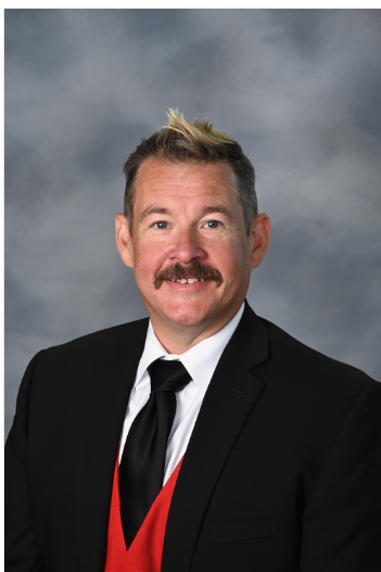 A man with short, styled hair, a mustache, wearing a black suit, white shirt, black tie, and red vest, posing in front of a gray background.