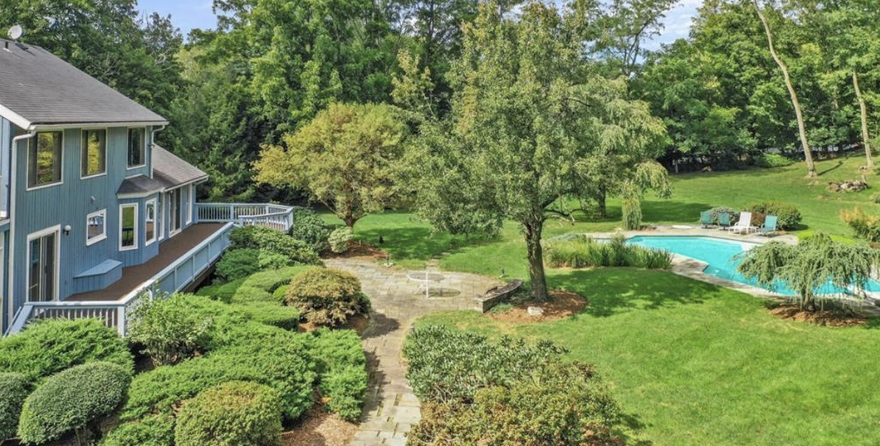 Backyard with a blue house on the left, lush green trees, a stone pathway, a swimming pool with lounge chairs around it, and well-maintained grass and shrubs.