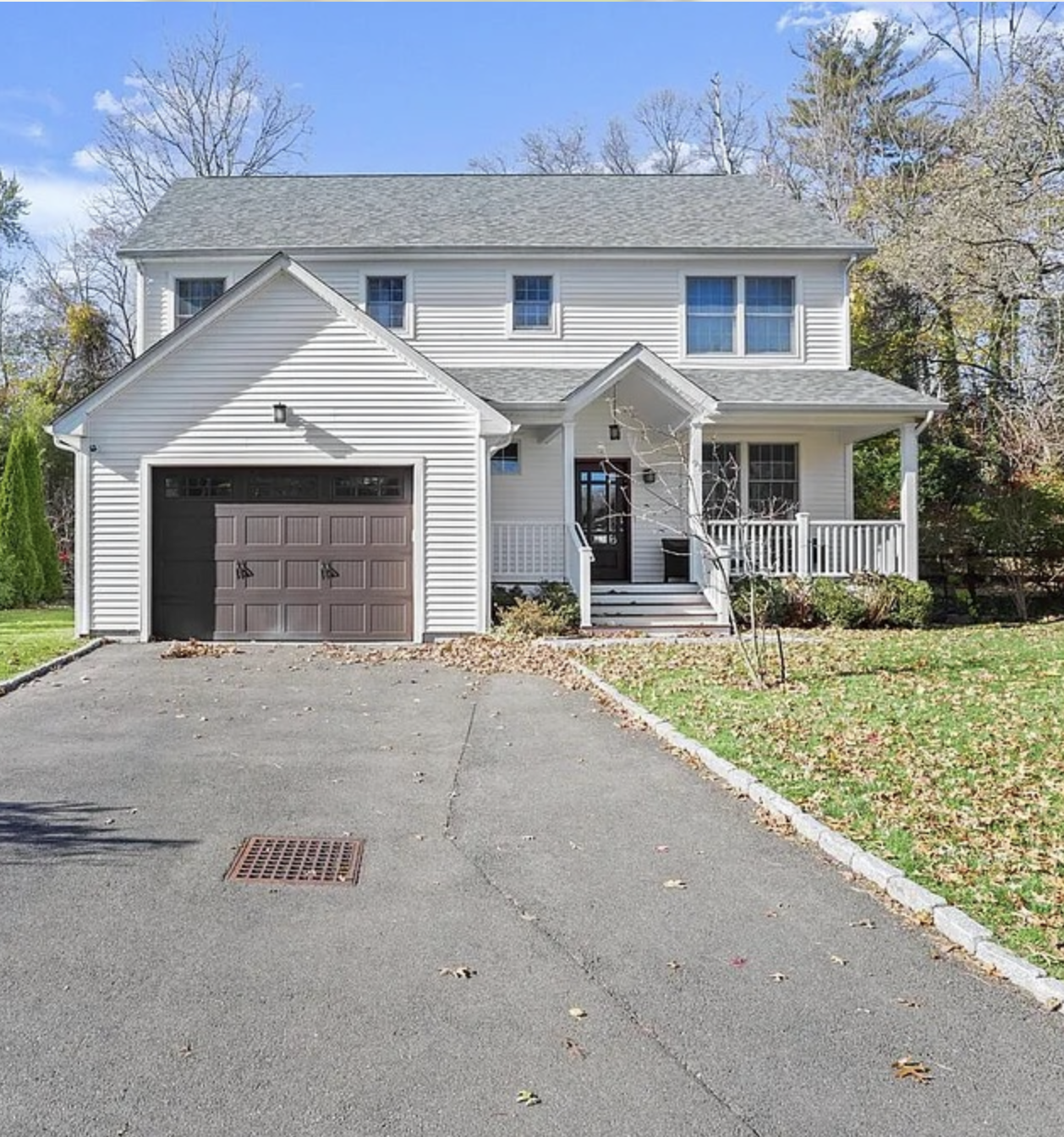 A two-story white house with a brown garage door and a front porch. The house has a gray shingled roof and several windows. The driveway is paved, and there are some fallen leaves on the grass and driveway. The scene is surrounded by trees and a partly cloudy sky.
