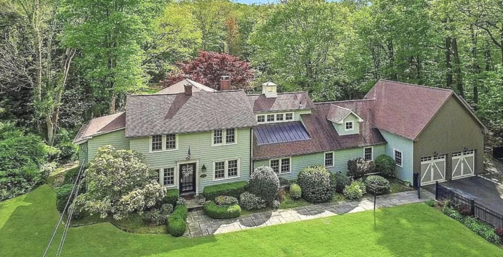 A large house painted light green surrounded by a well-manicured lawn, bushes, and flowering trees. The house has a brown shingle roof, several chimneys, and a detached garage with white doors. A stone pathway leads to the front door, and trees and forested area are visible in the background.