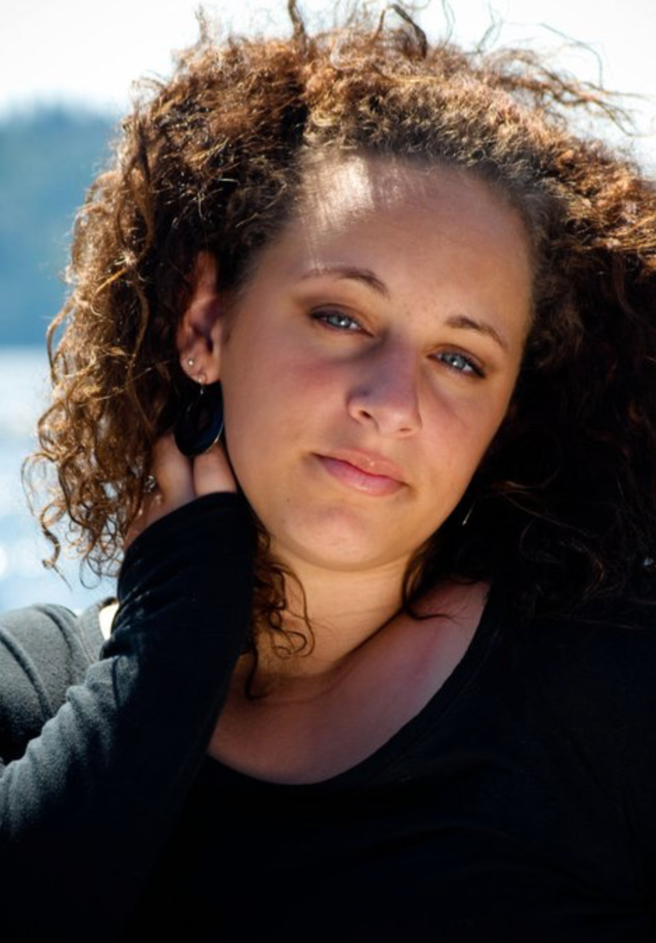 Close-up portrait of a woman with curly hair, blue eyes, and earrings, outdoors with sunlight.