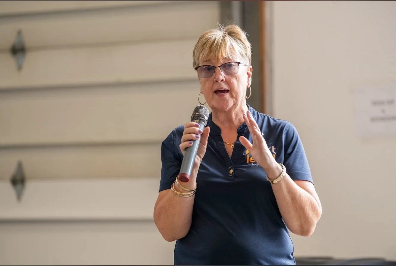A woman with short blonde hair, wearing glasses and a navy blue shirt, speaking into a microphone indoors.
