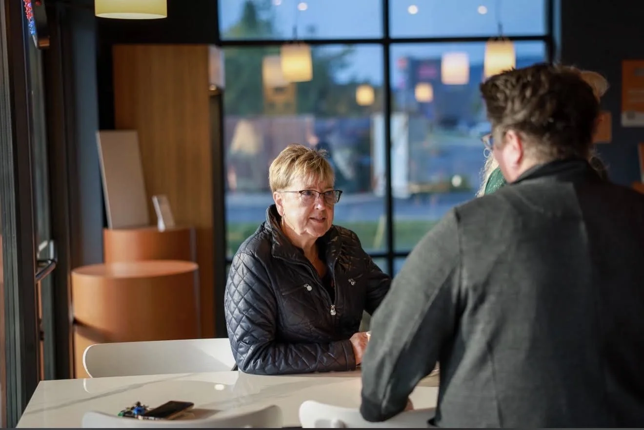 An older woman with glasses and short hair, wearing a black quilted jacket, talking to two younger people, one of whom has dark hair and glasses, seated at a white table in a modern, well-lit café with large windows.