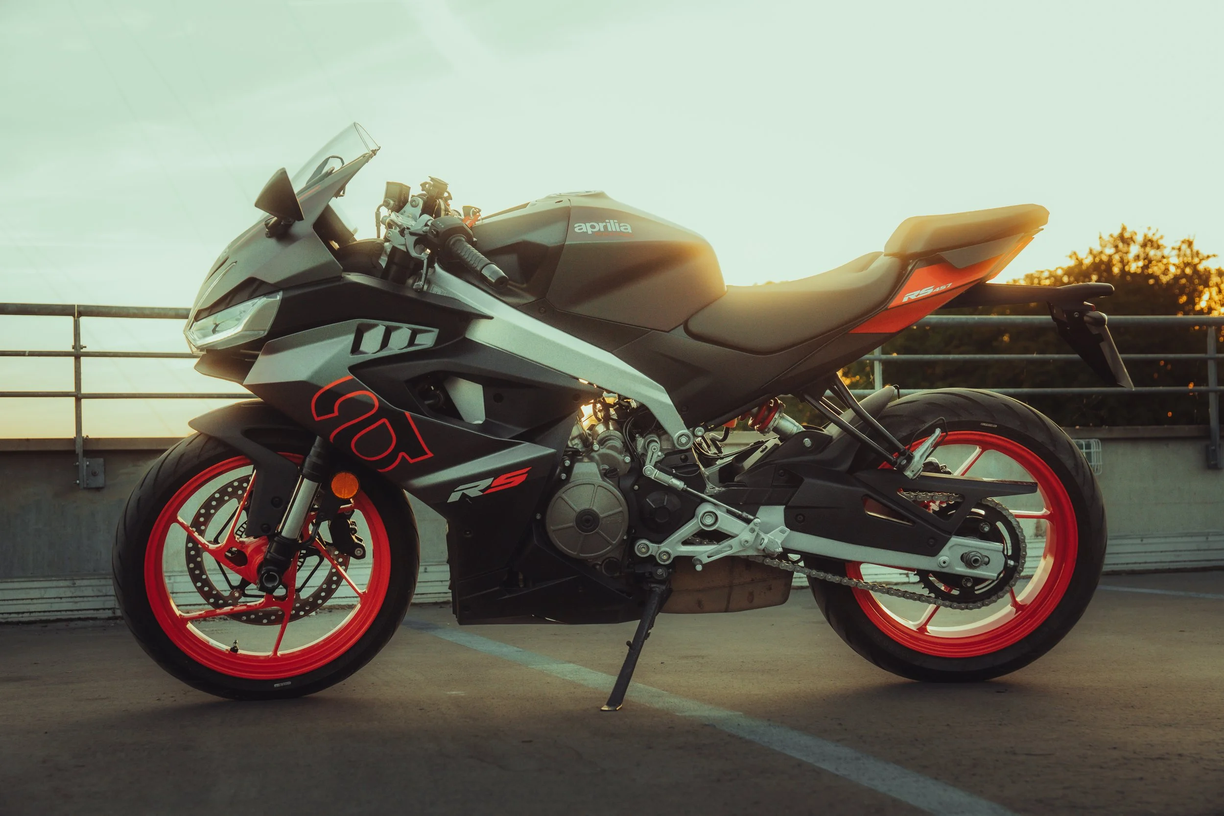 Une moto de sport noire avec des accents rouges, marquée 'Aprilia', stationnée dans une zone de parking, avec une barrière en arrière-plan et un ciel ensoleillé au crépuscule.