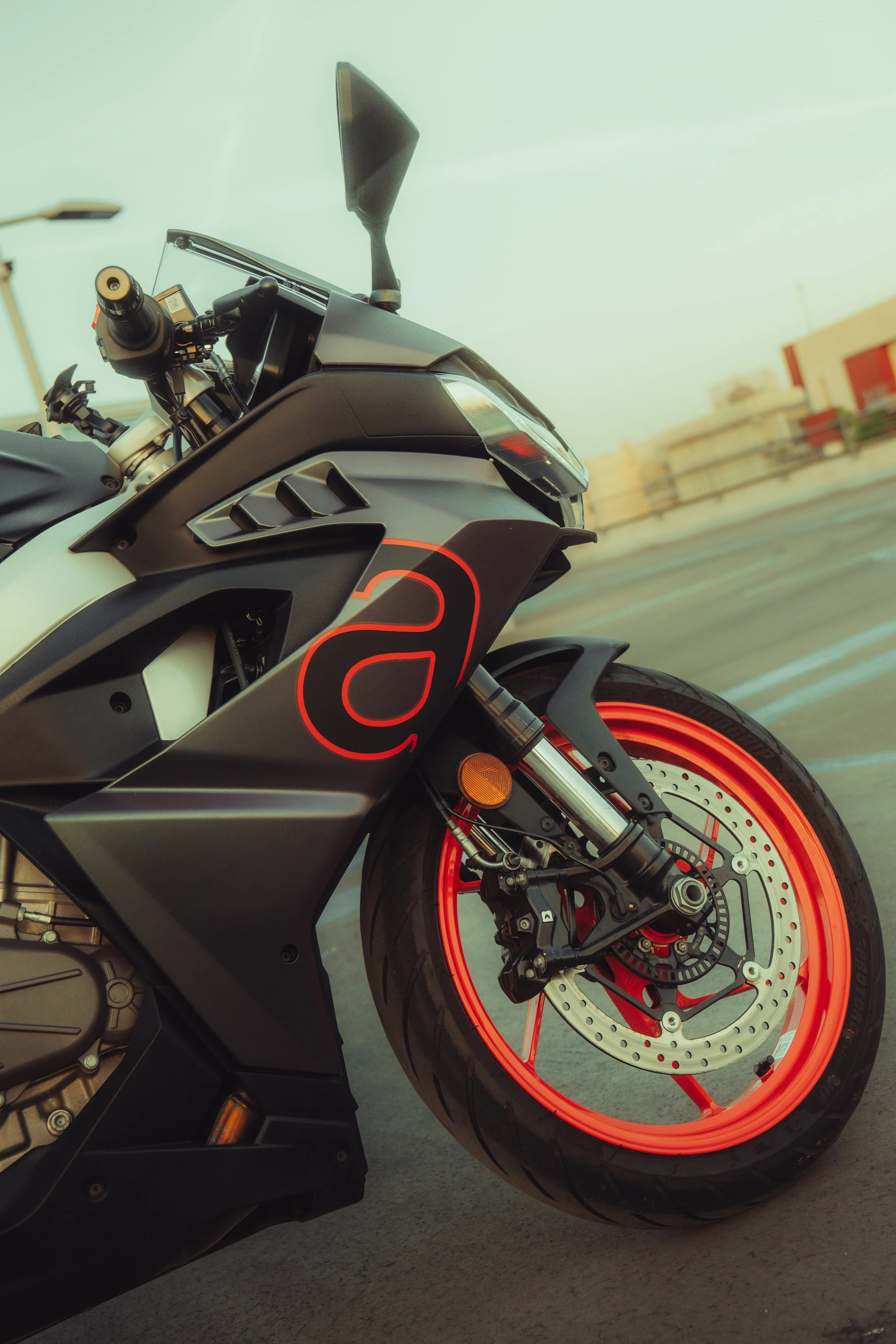 Une moto noire avec des accents orange, vue de côté, stationnée sur une route avec des bâtiments flous en arrière-plan.
