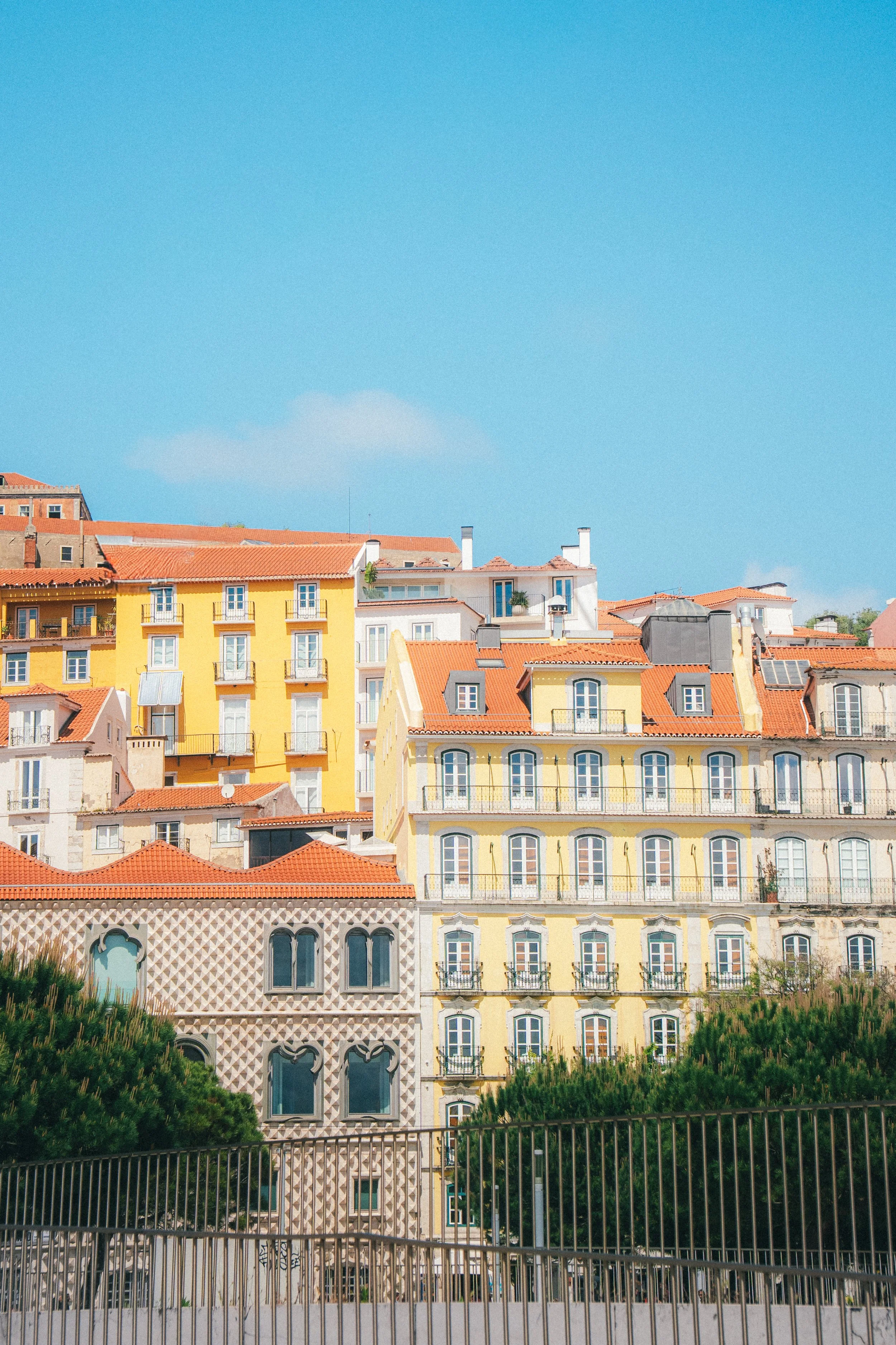 Un ensemble de bâtiments résidentiels colorés avec des toits en tuiles rouges, vue à travers une grille noire en métal, sous un ciel bleu clair.