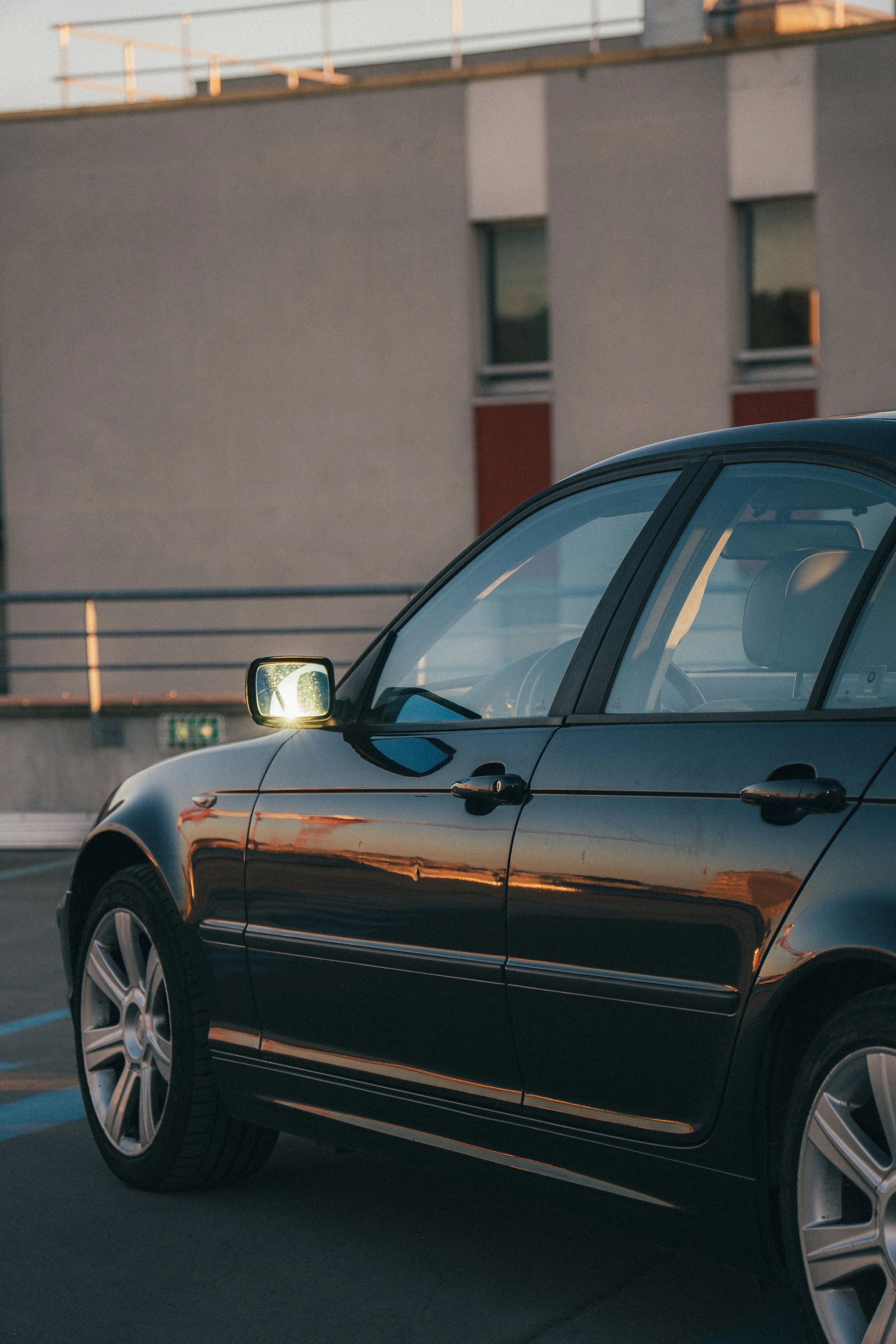Vue partielle d'une voiture noire garée dans un parking avec un bâtiment moderne en arrière-plan, éclairé par la lumière du coucher de soleil.