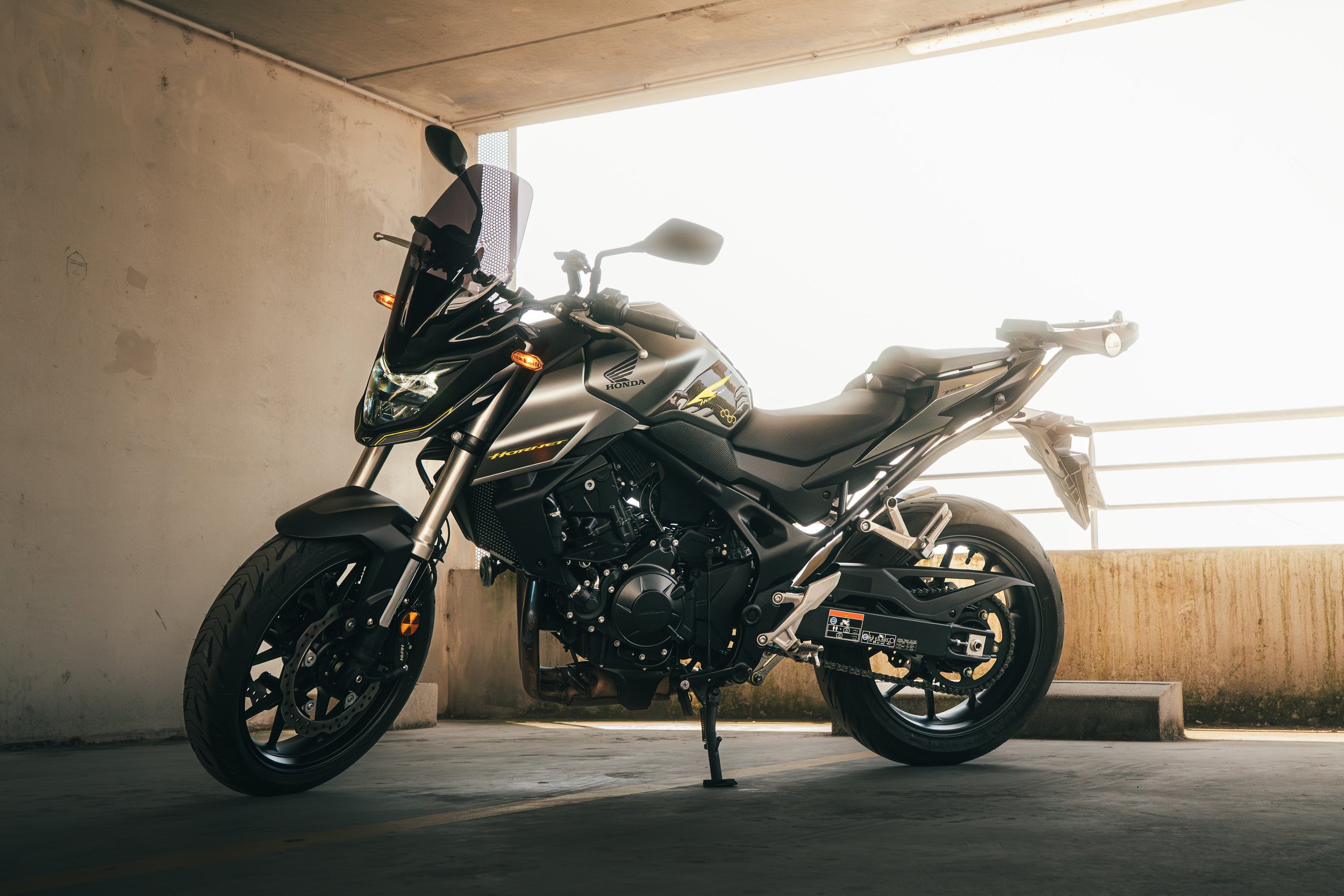Une moto Honda noire, stationnée dans un parking sous un plafond en béton, éclairée par une lumière vive à l'arrière.