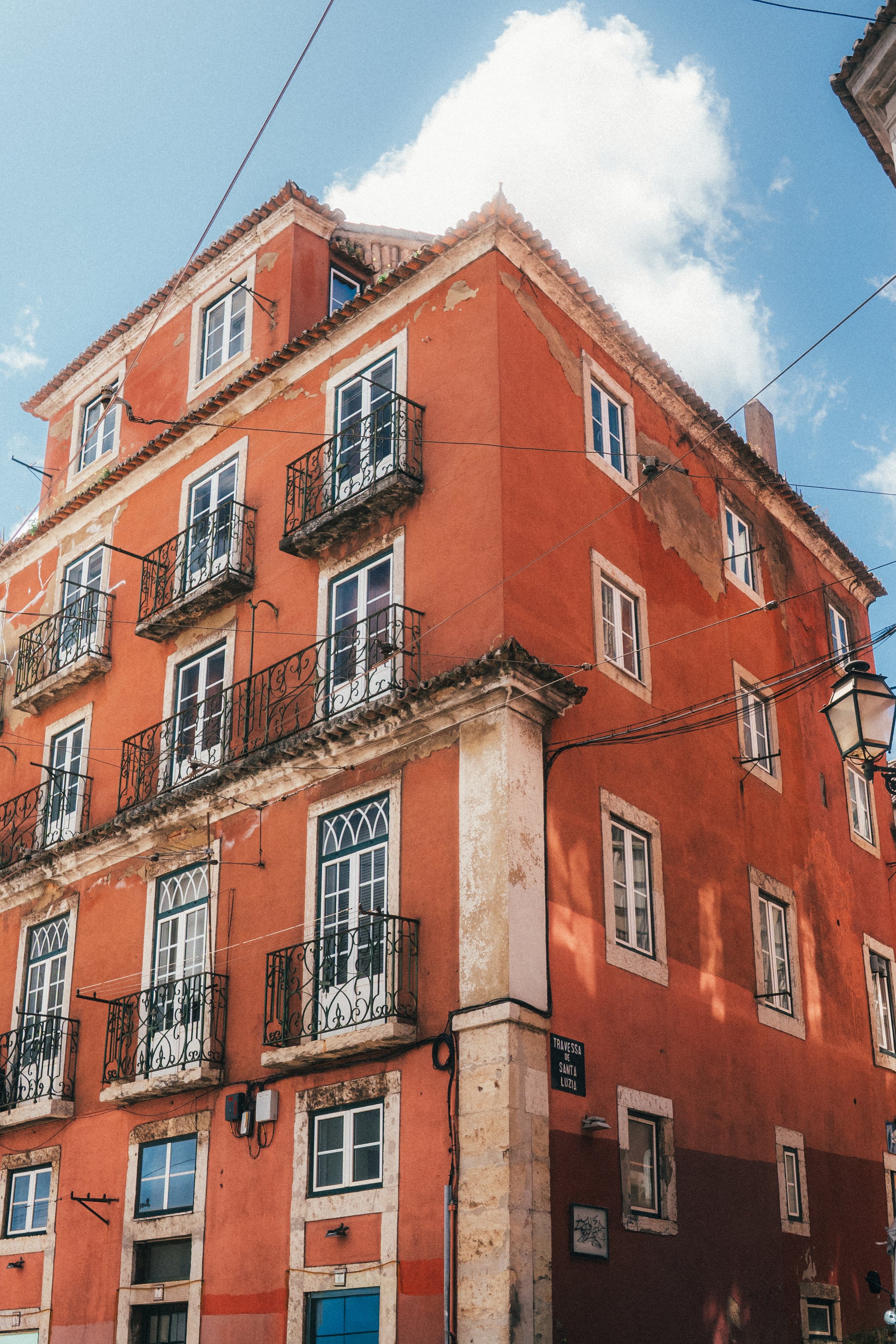 Un bâtiment résidentiel orange avec plusieurs fenêtres à volets blancs et balcons en fer forgé, situé dans une zone urbaine avec un ciel bleu et quelques nuages, rue Travessa de Santa Luzia.