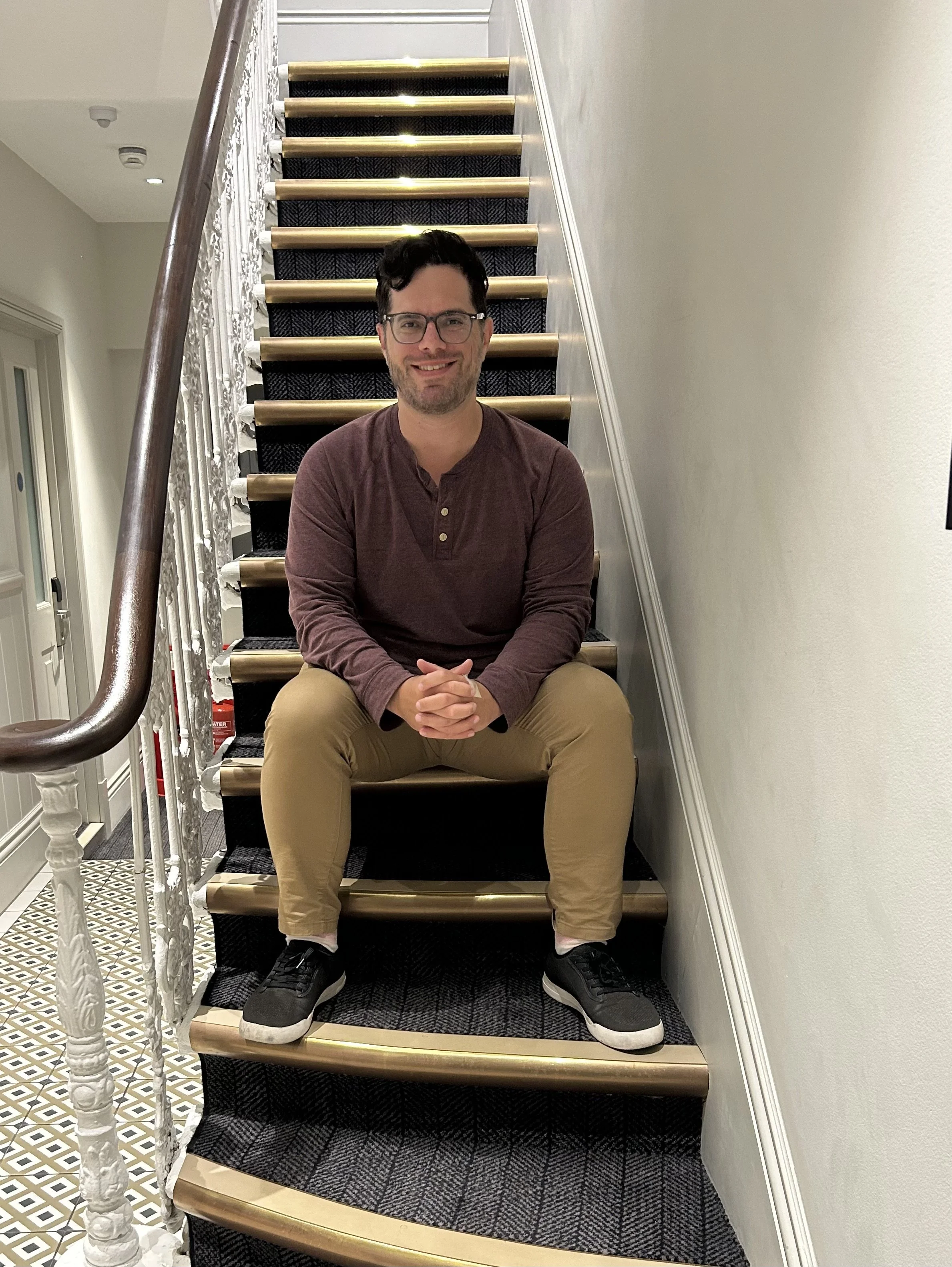 Man sitting on staircase with black sneakers, beige pants, maroon shirt, glasses, and dark hair, smiling in indoor setting.