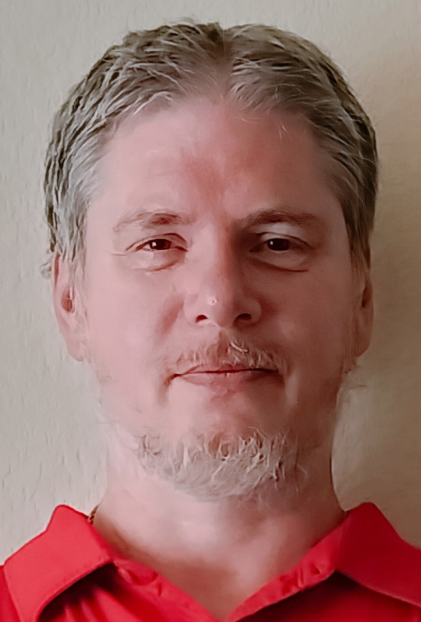 A middle-aged man with gray hair and beard, wearing a red shirt, smiling gently, standing against a beige wall.