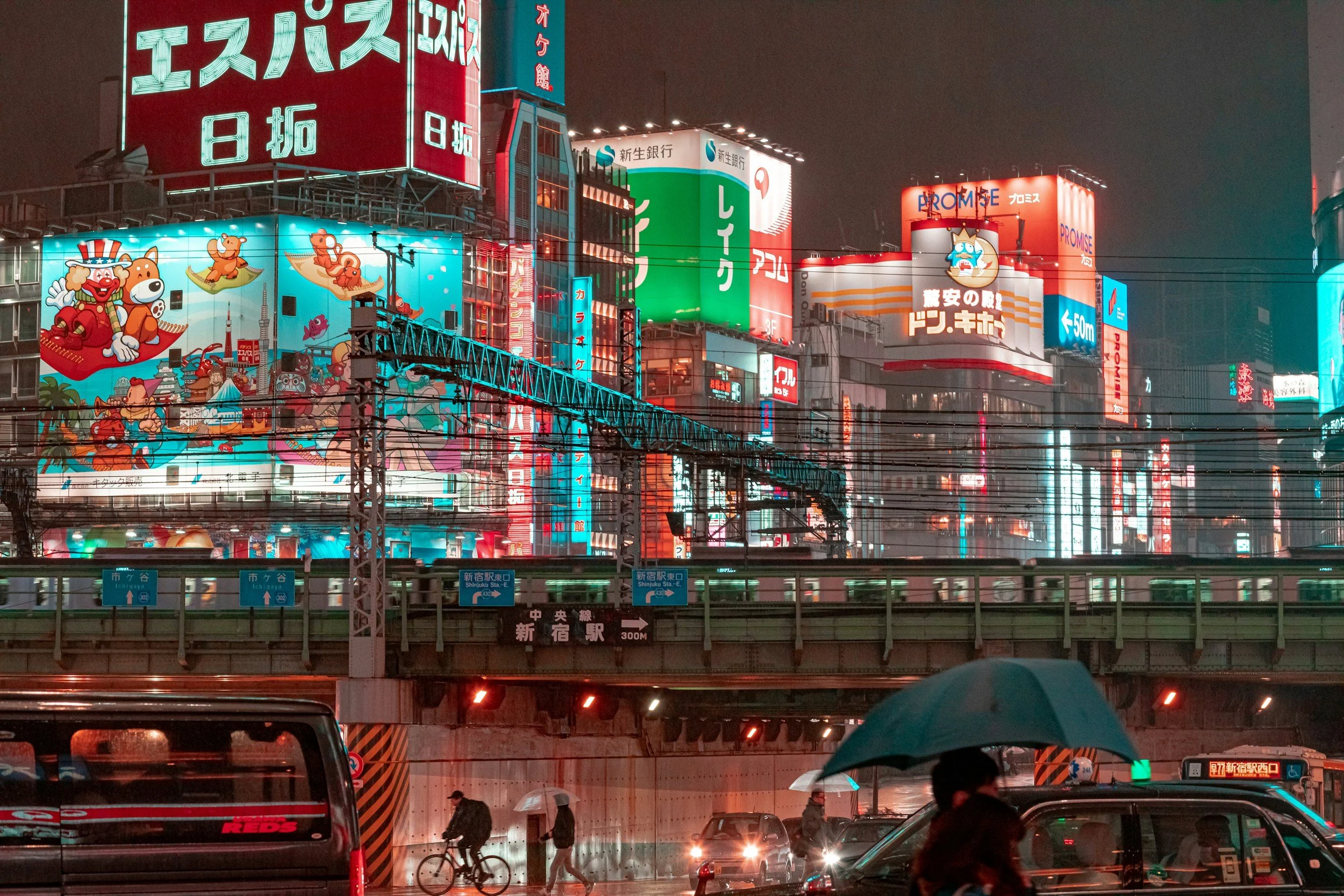 A city street scene at night with illuminated advertisements and signs in Japanese. In the foreground, people with umbrellas walk and cars are parked. Above, multiple large colorful billboards display Japanese text and cartoon characters, including a bear and other animals, advertising various businesses.