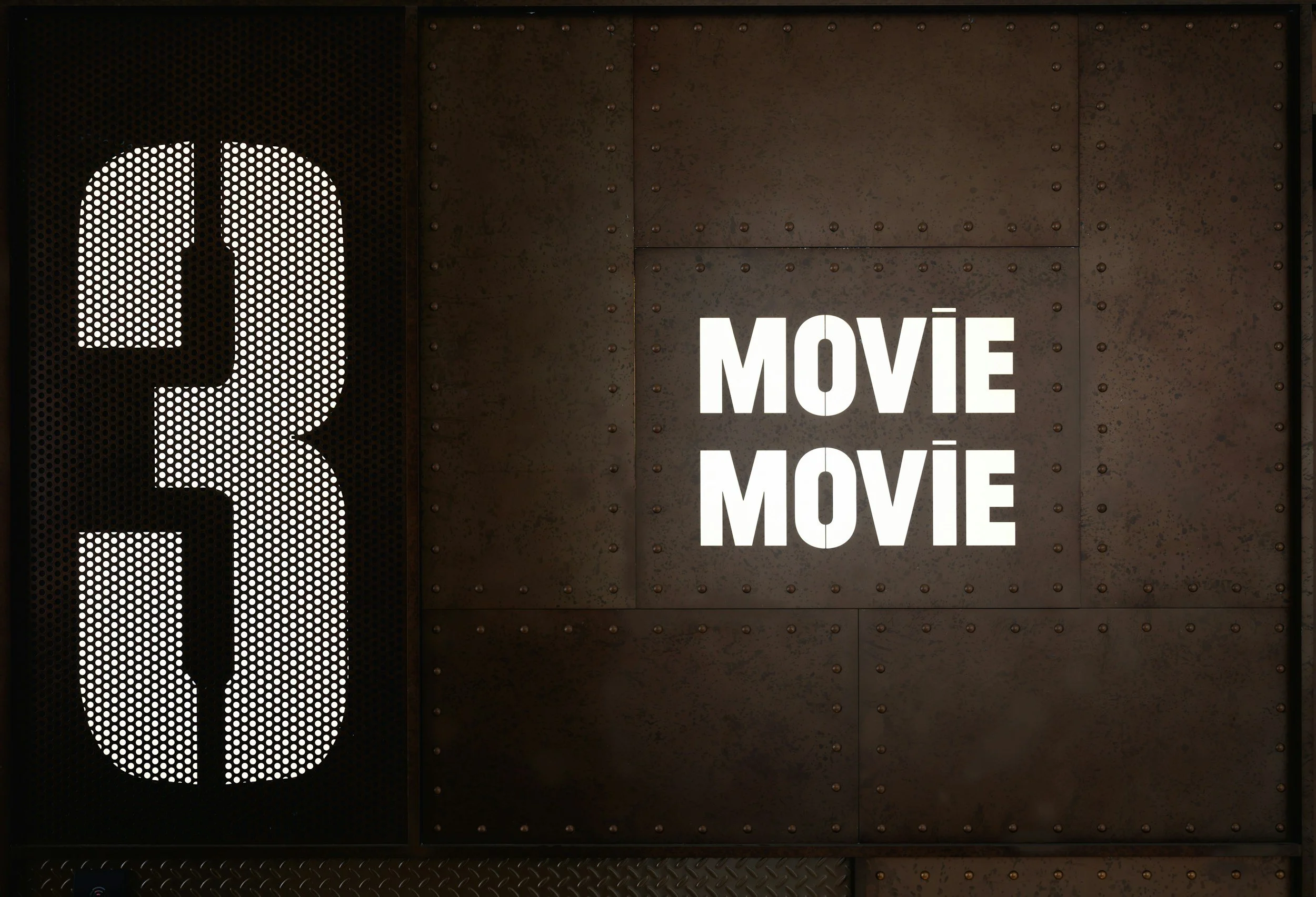An entrance for a movie theater with a large illuminated sign that says 'MOVIE' in white capital letters. The door features a stylized film reel icon created with a pattern of white dots.