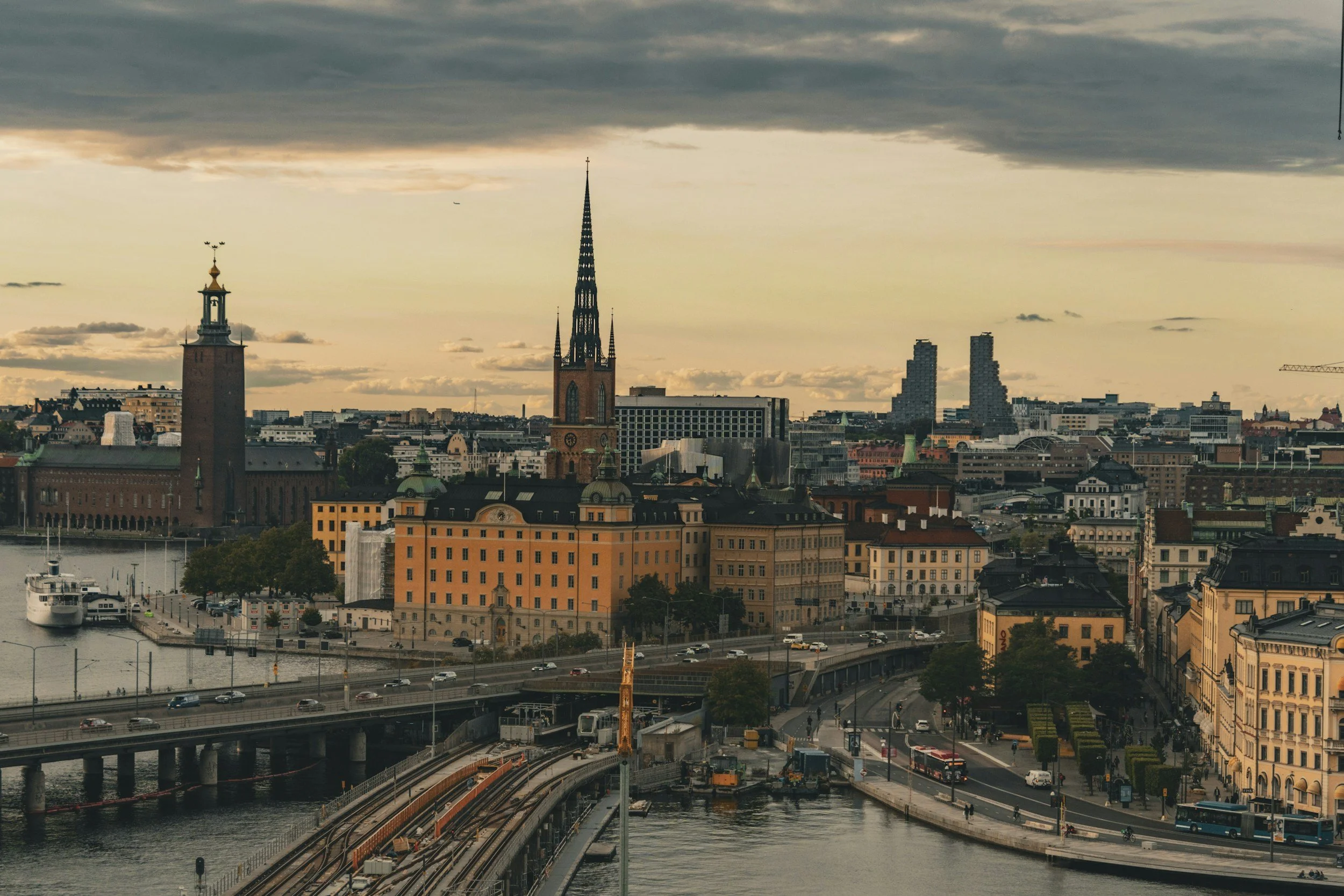 City skyline featuring historic and modern buildings, a church with a tall spire, a waterfront with a docked boat, and traffic on a bridge, under a cloudy sky.
