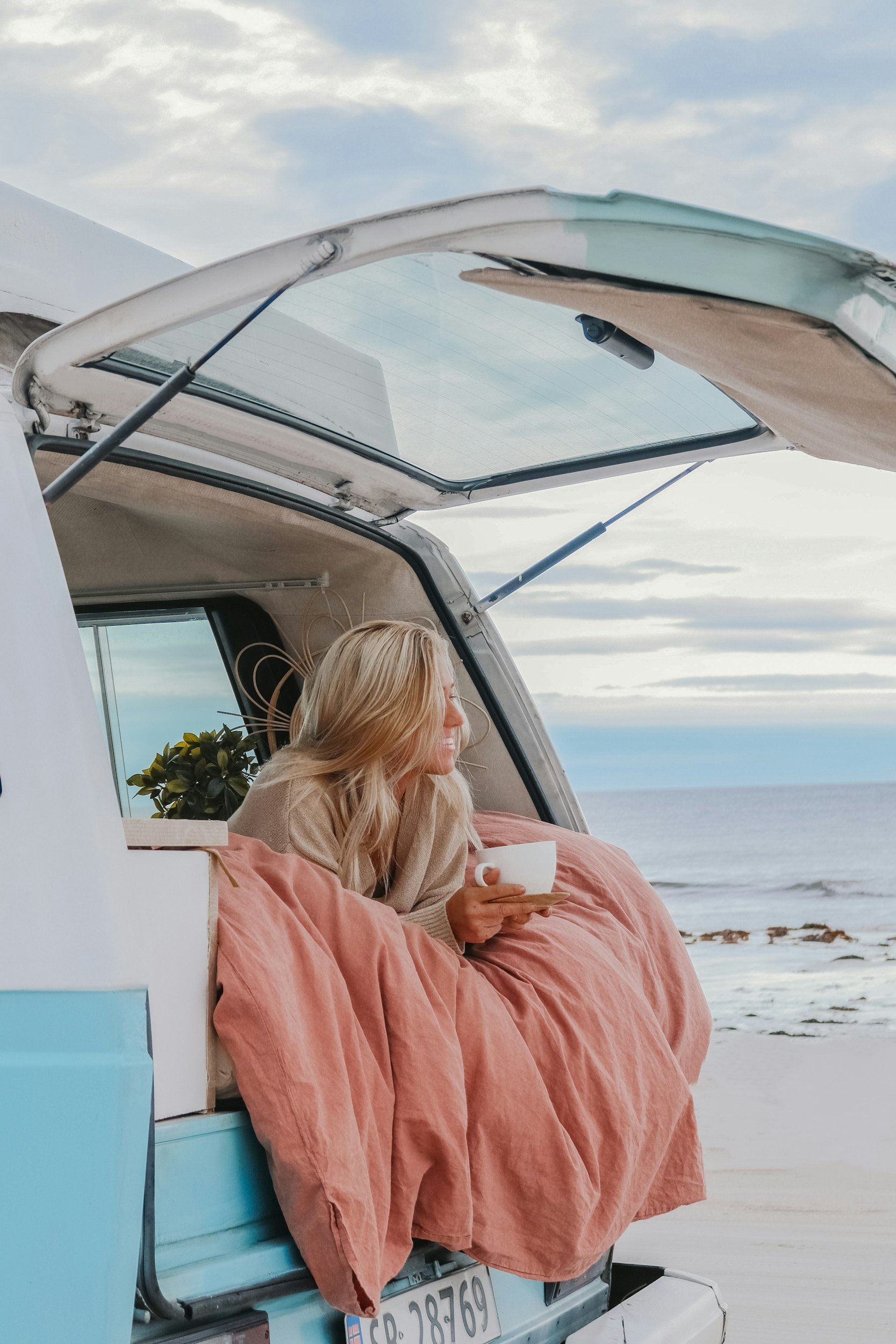 A woman with blonde hair relaxing in a boat or camper at the beach, holding a white mug, with a view of the ocean and cloudy sky in the background.