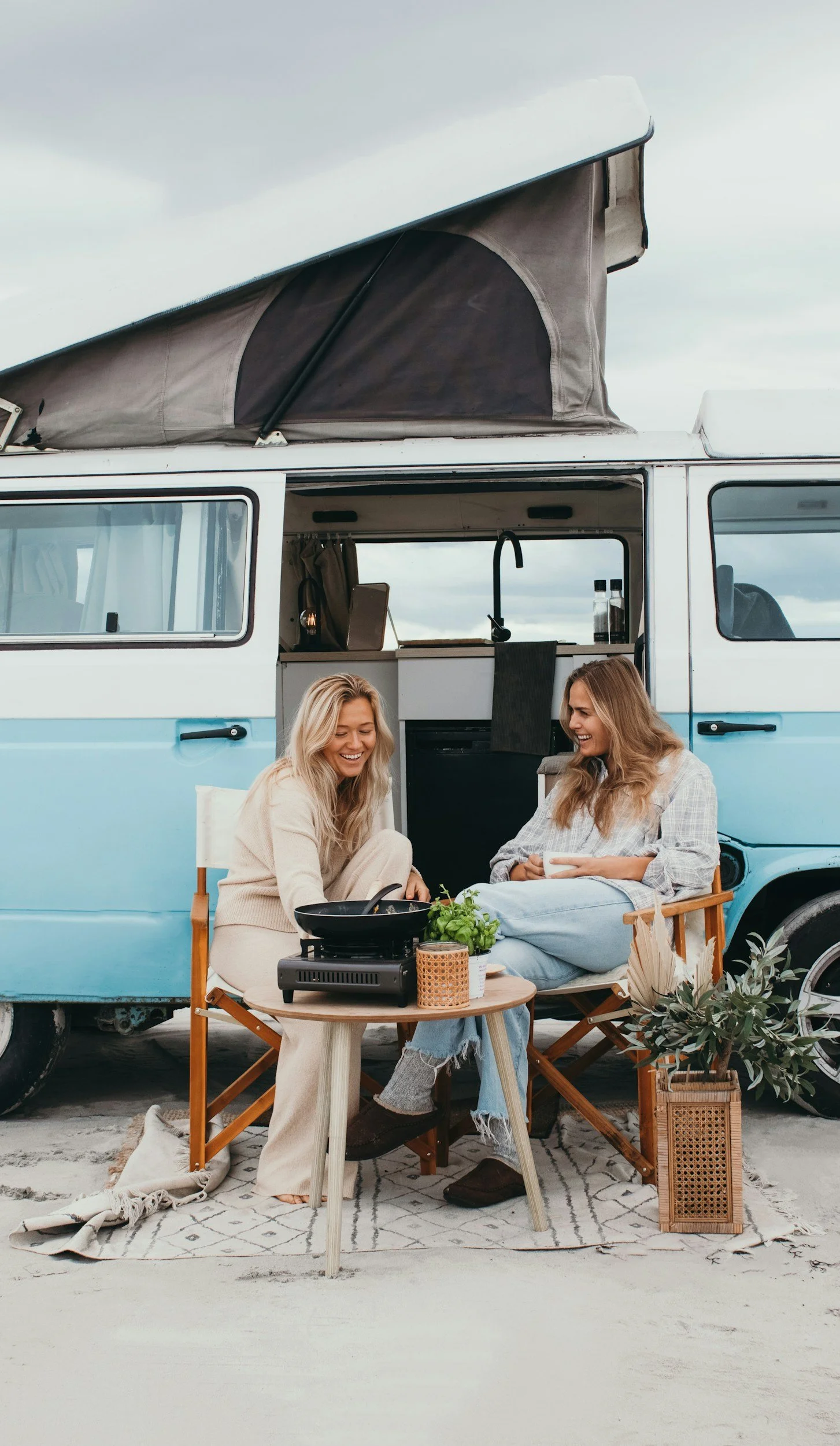 Two women sitting outside a vintage van with an extended rooftop camper, enjoying a meal together with a portable stove, plants, and cozy decor, under a cloudy sky.