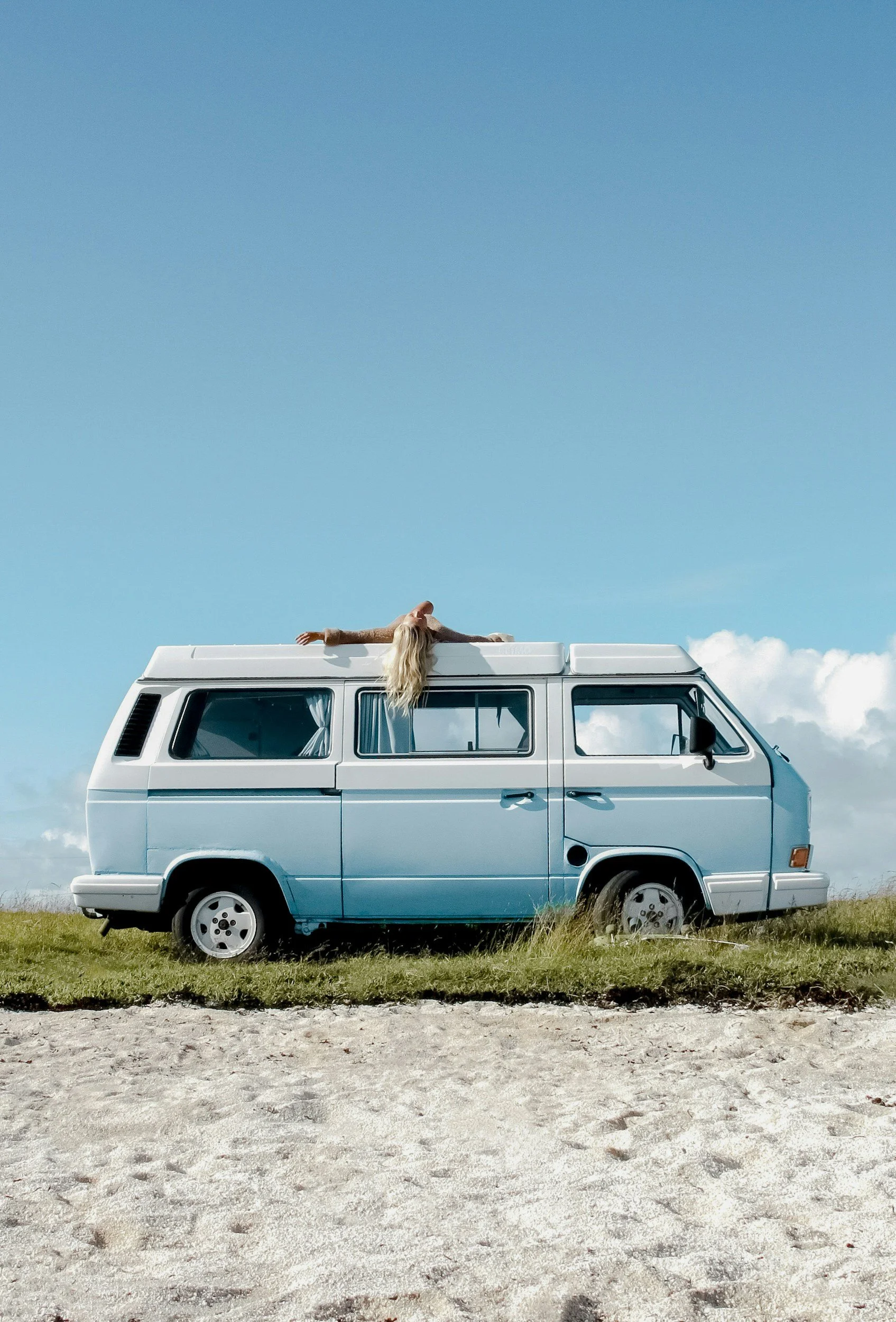 A woman with long blonde hair lying on top of a light blue vintage van with her arms outstretched, against a bright blue sky with a few clouds. The van is parked on a grassy area with sand in the foreground.