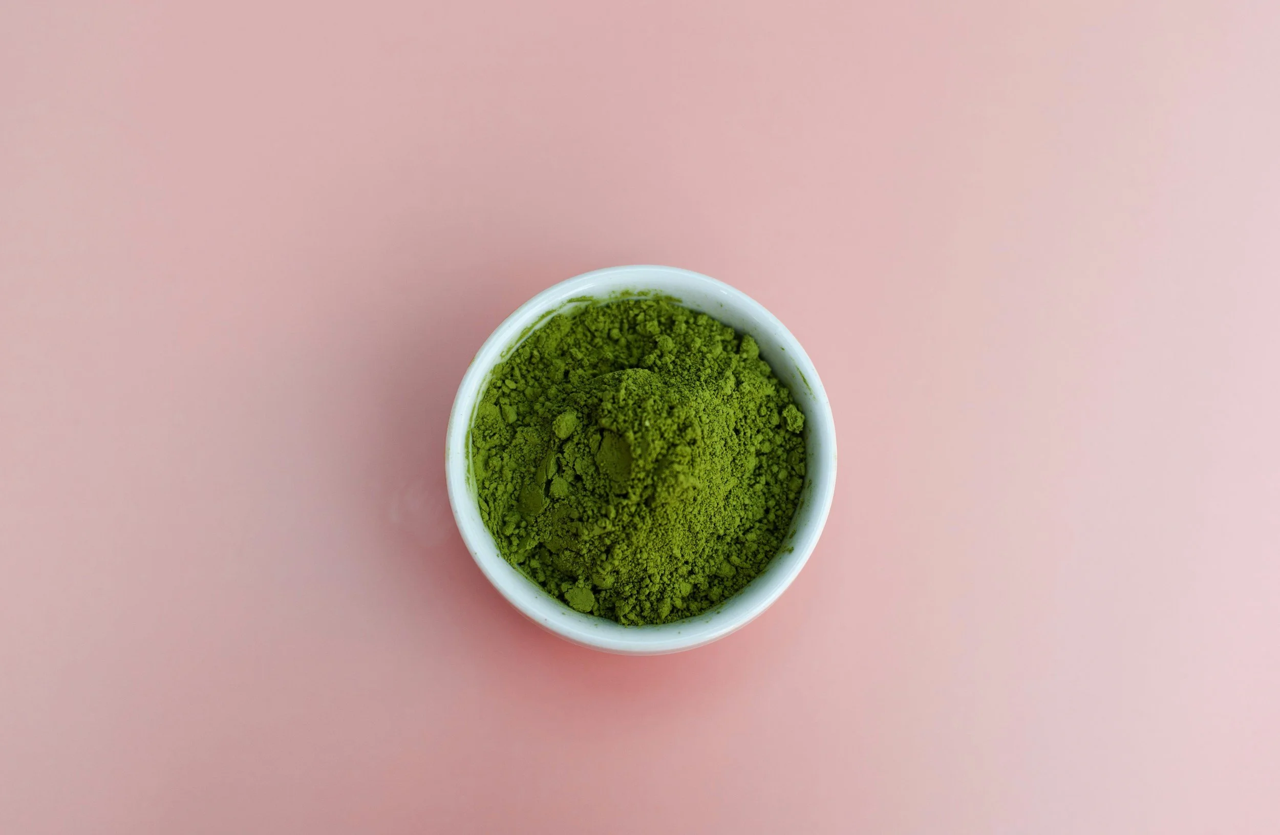 Close-up of a small white bowl filled with green powder on a pink background.