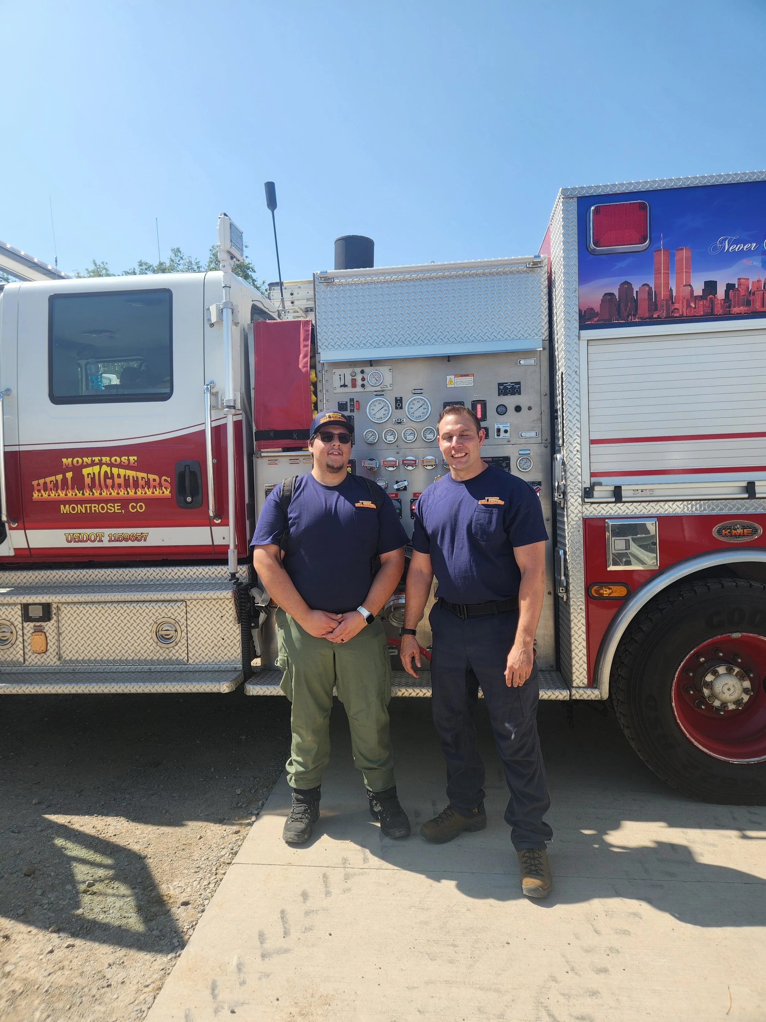 Two firefighters stand in front of a fire truck, smiling at the camera during daytime with a clear blue sky.