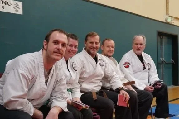 Five men in martial arts gi uniforms sitting on a bench in a dojo.
