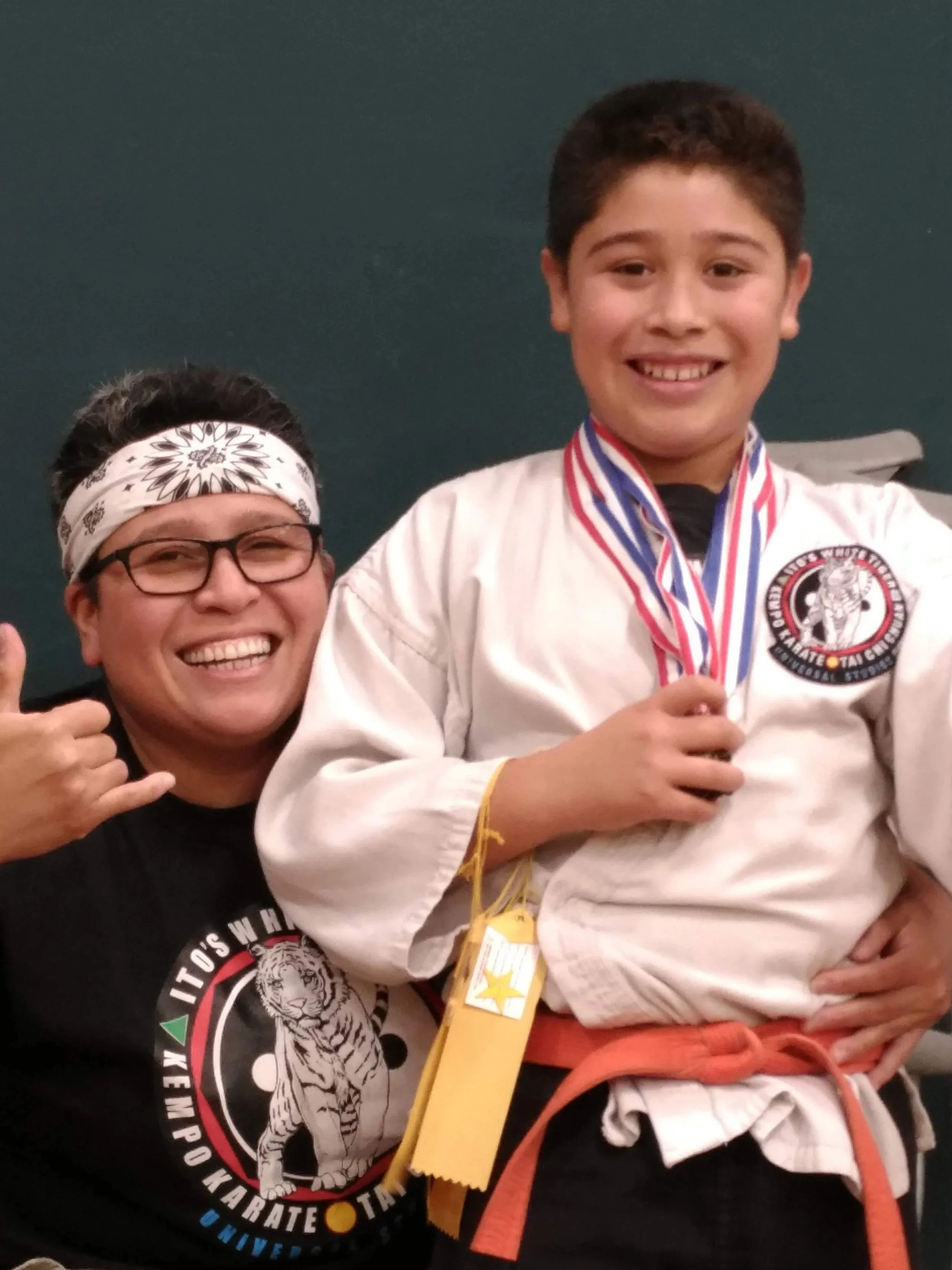 Young boy in a taekwondo uniform with a medal, standing next to a smiling man wearing glasses and a bandana, both celebrating their achievement.
