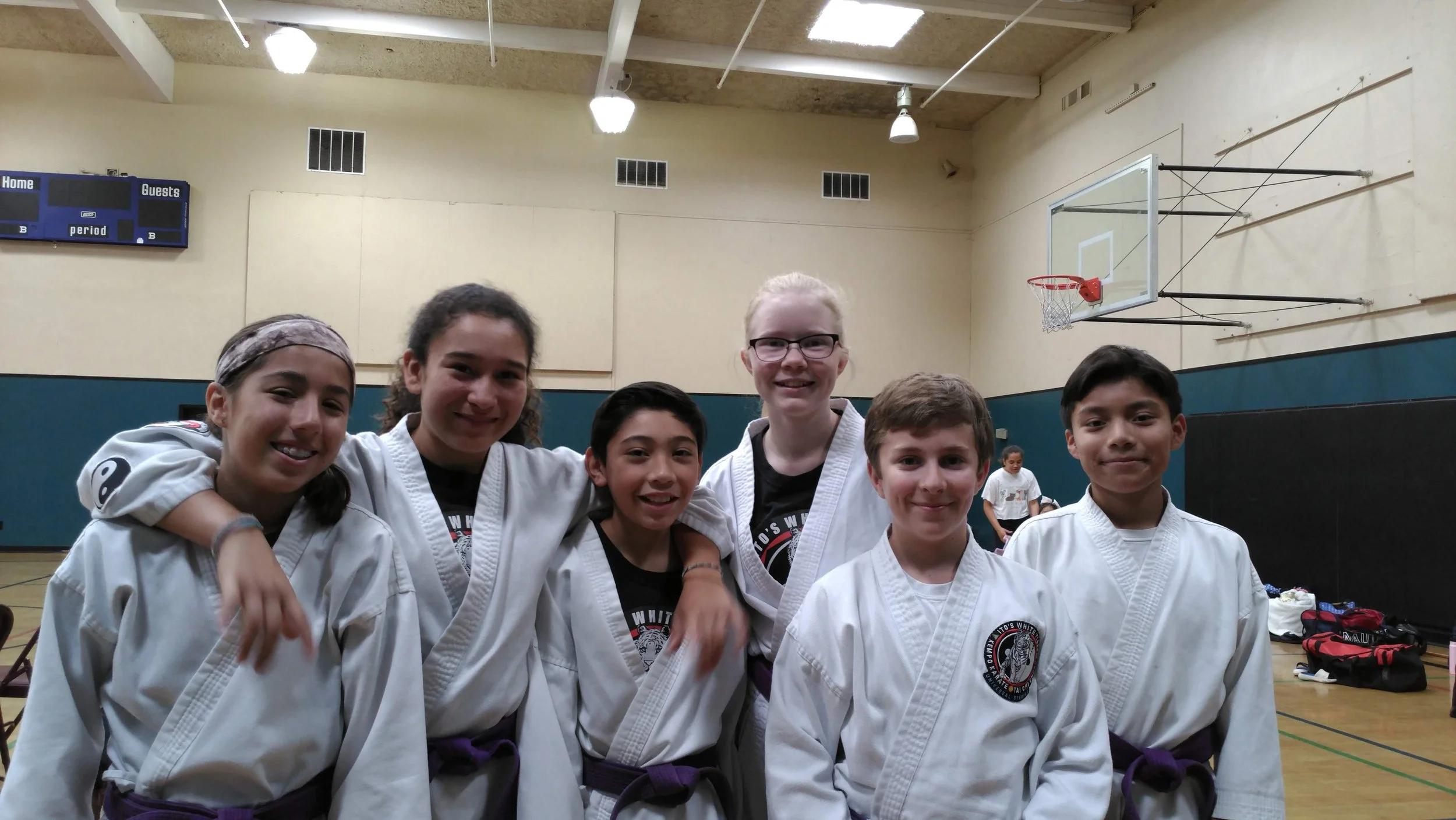 Group of seven children in martial arts uniforms smiling at the camera in a gymnasium.
