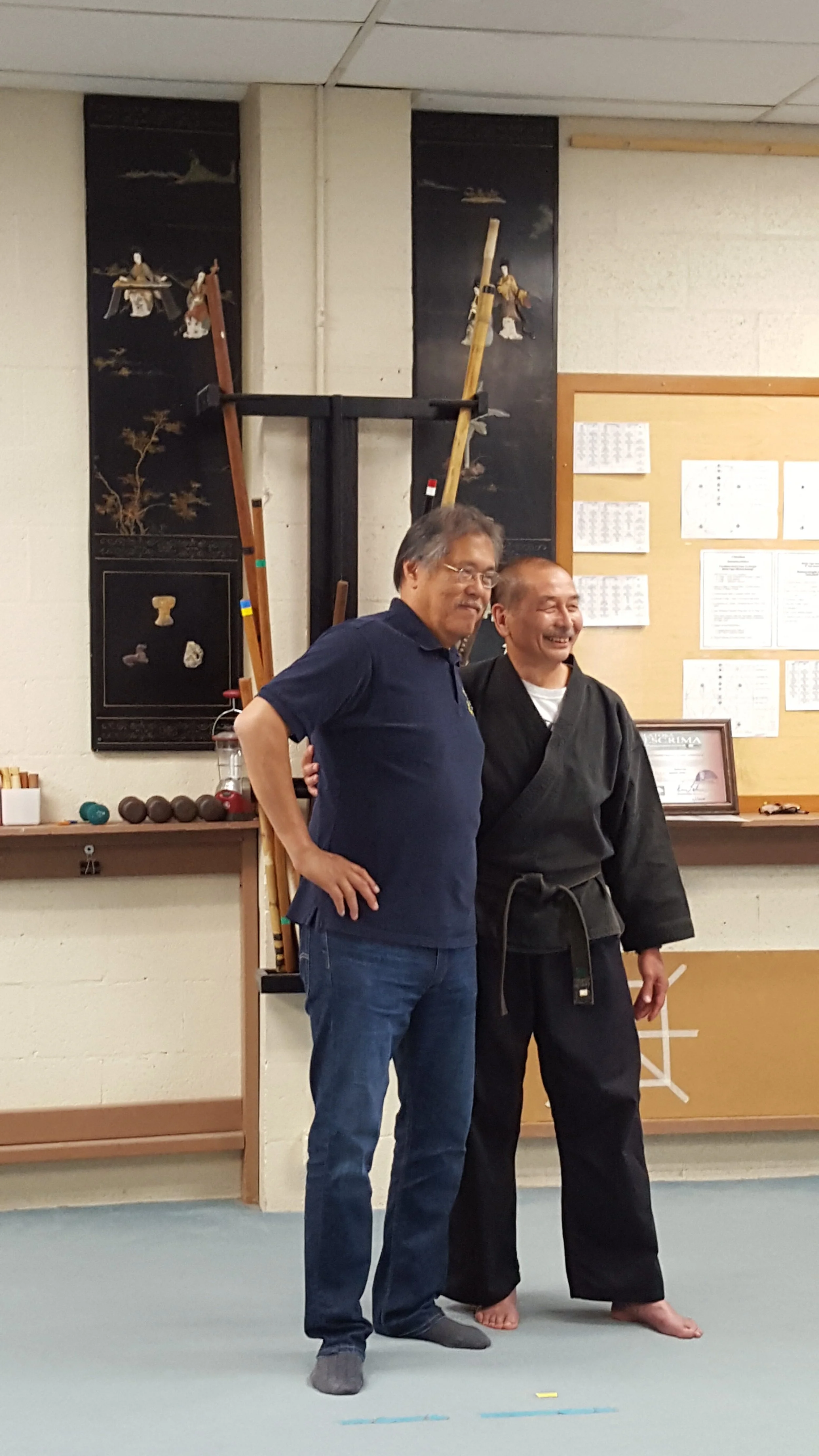 Two men standing together indoors, one in casual clothing and the other in a martial arts uniform. They are smiling and posing for the photo.