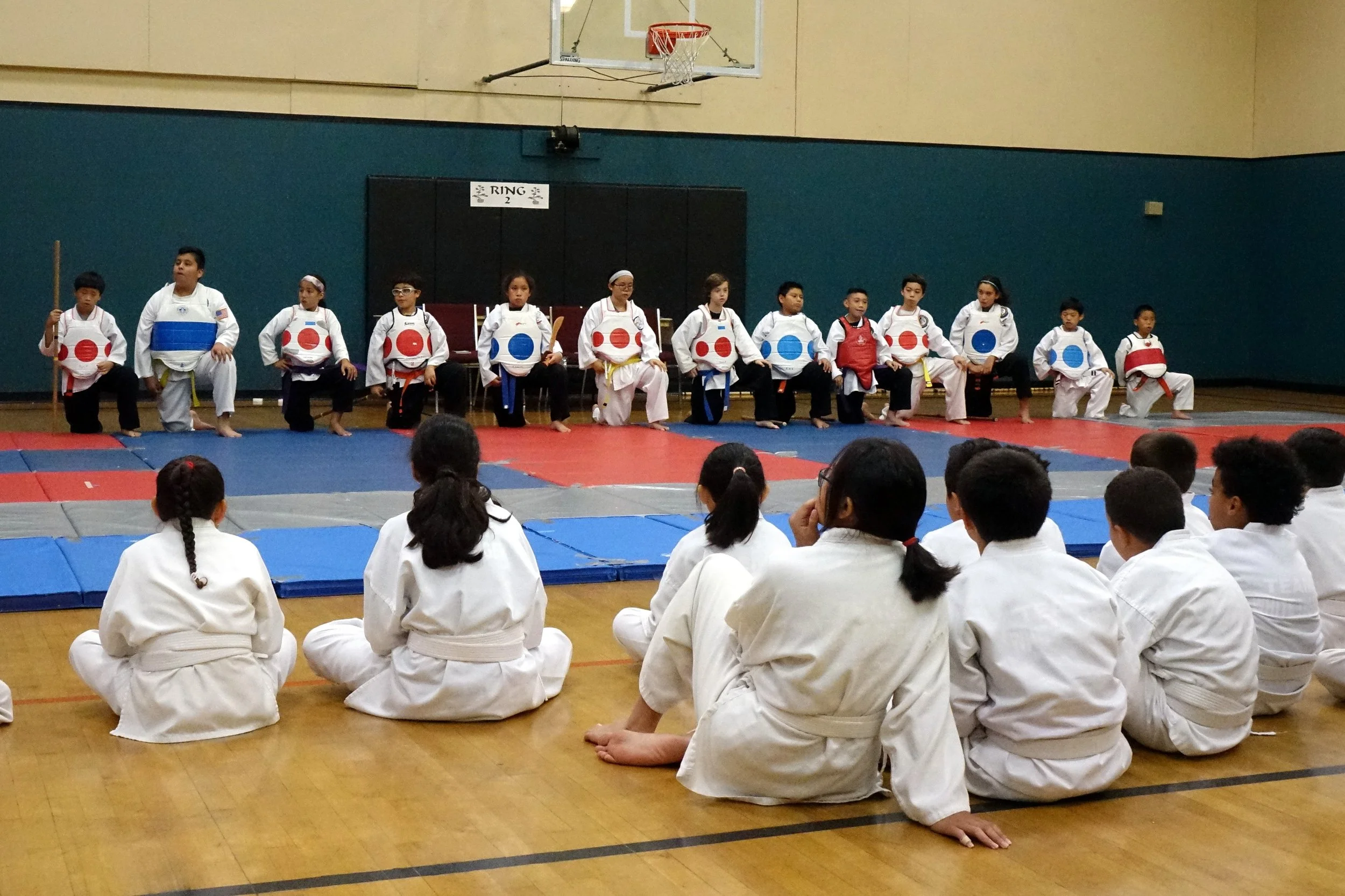 Children in martial arts uniforms sitting on the floor and kneeling in front of a stage with students in martial arts uniforms performing in front of an audience in a gymnasium.