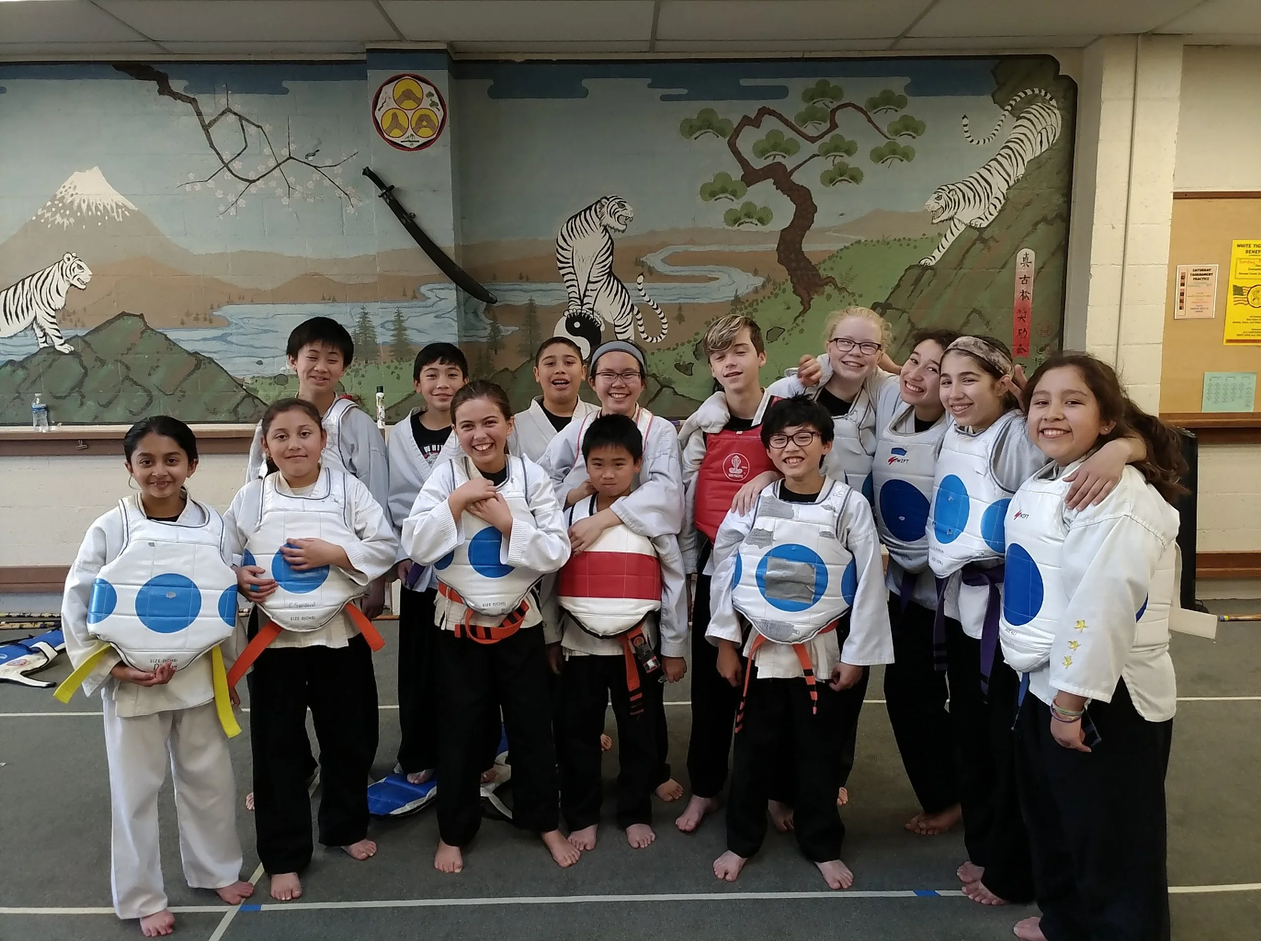 Group of children in martial arts uniforms, smiling and posing in front of a mural depicting tigers, Mount Fuji, cherry blossoms, and nature scenes.