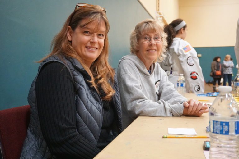 Two women sitting at a table with water bottles, with a group of people and children in the background.