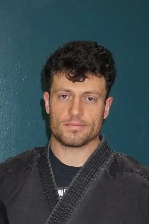 A young man with short curly dark hair, light skin, and facial hair, wearing a black martial arts gi with a black belt, standing in front of a dark teal wall.